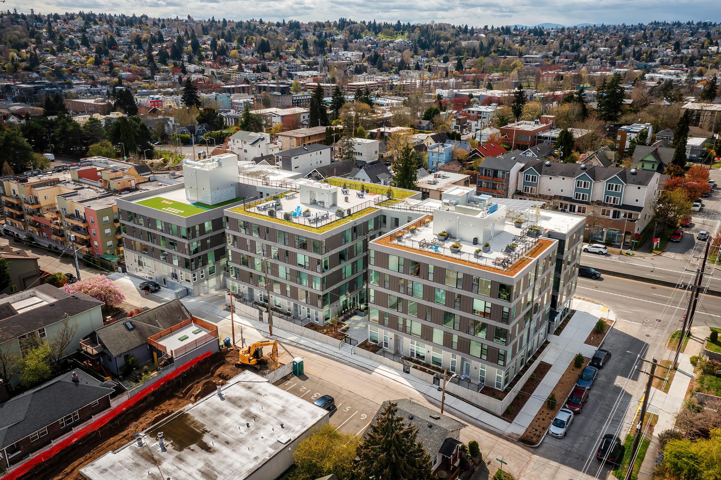 Aerial view of modern residential buildings with flat roofs and green spaces on top, surrounded by a mix of urban houses and greenery. The scene includes streets, trees, and a distant skyline under a partly cloudy sky, showcasing a vibrant neighborhood setting.