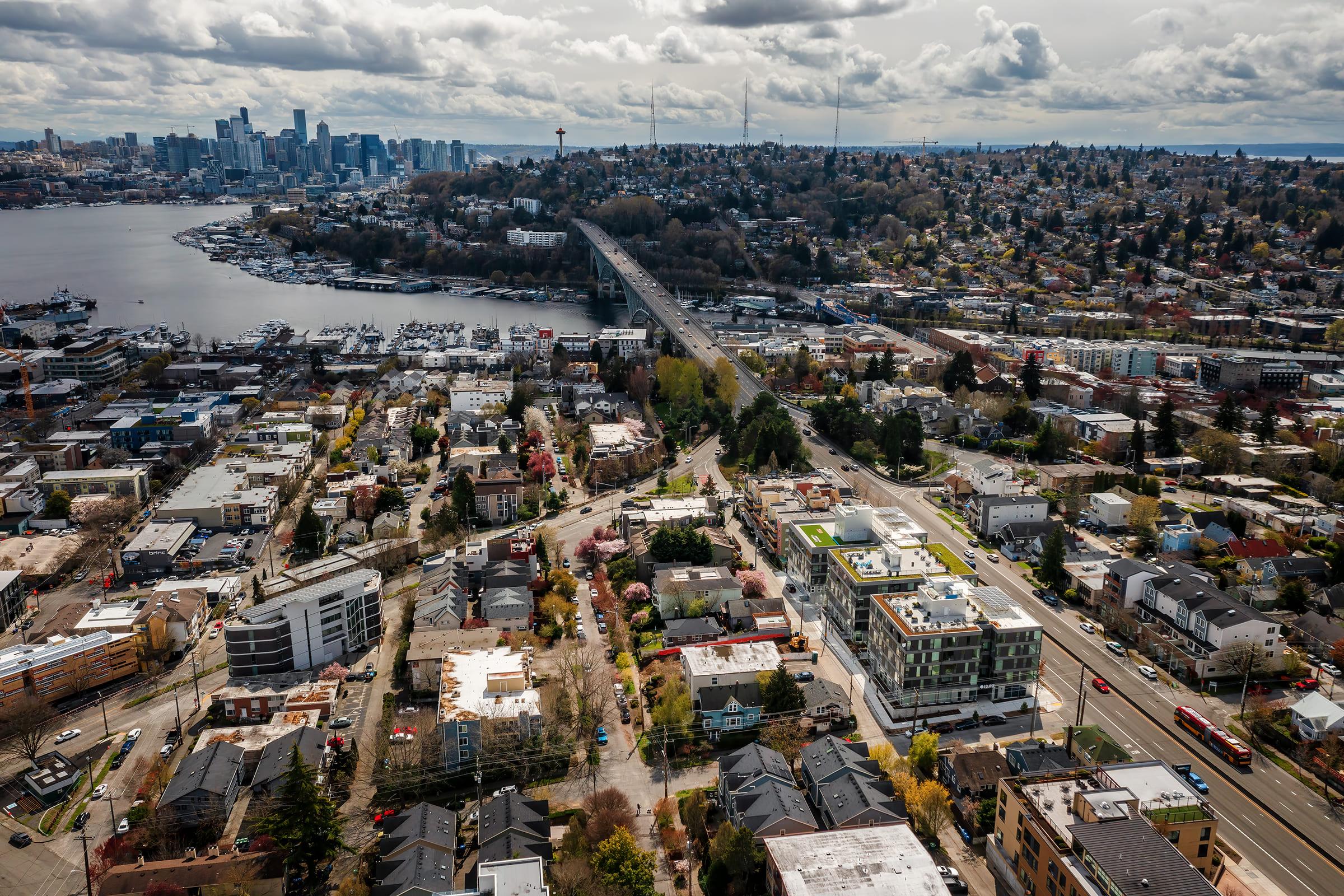 Aerial view of Seattle showcasing the waterfront, the Space Needle, and various neighborhoods. The scene includes a mix of residential and commercial buildings, with a backdrop of the city skyline and clouds overhead. The image captures the vibrant urban landscape and surrounding natural beauty.