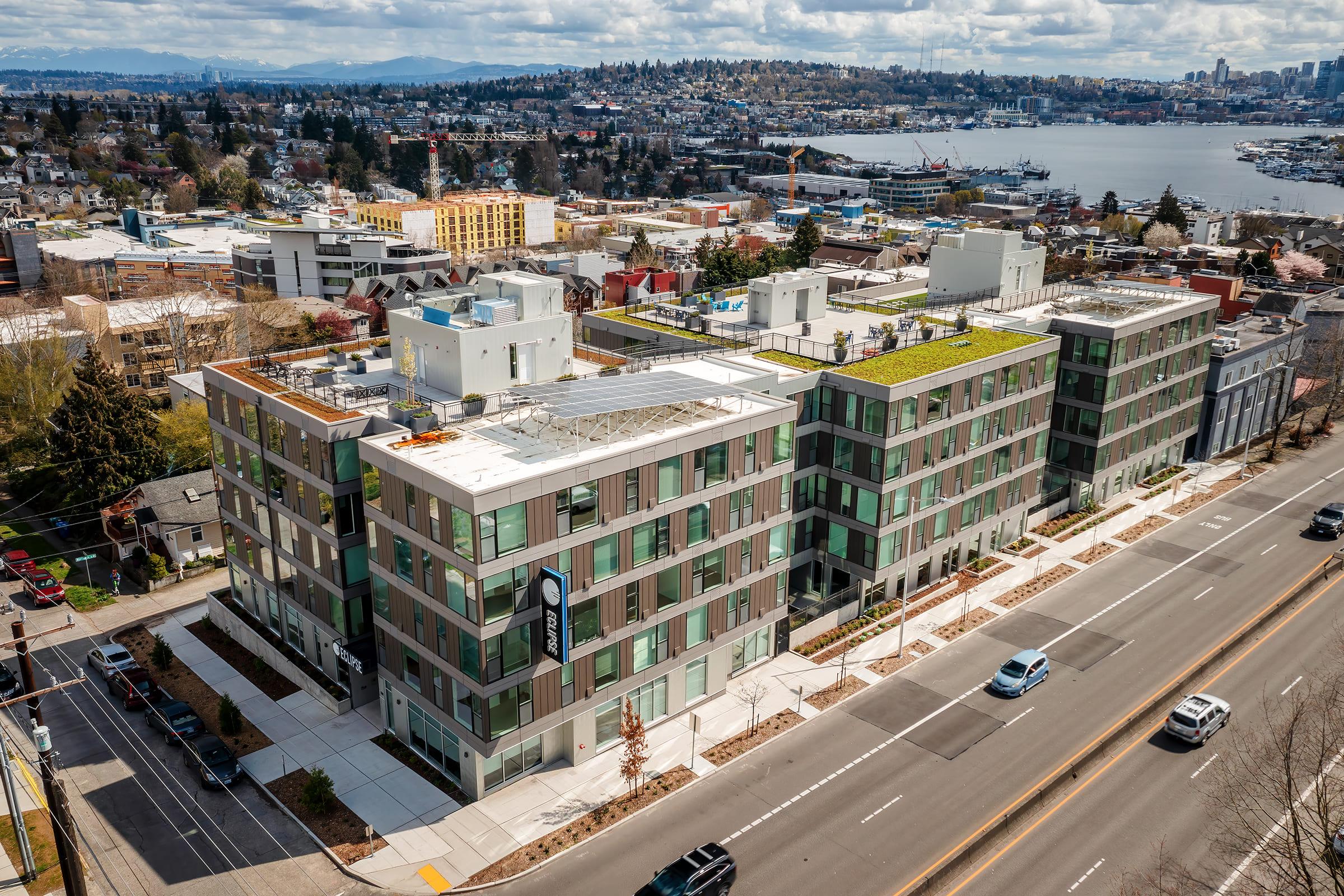 Aerial view of modern multi-story buildings along a street, featuring green roofs and large windows. The background includes a scenic landscape with hills and a body of water, while the foreground shows well-maintained sidewalks and landscaping. The setting appears urban and vibrant.