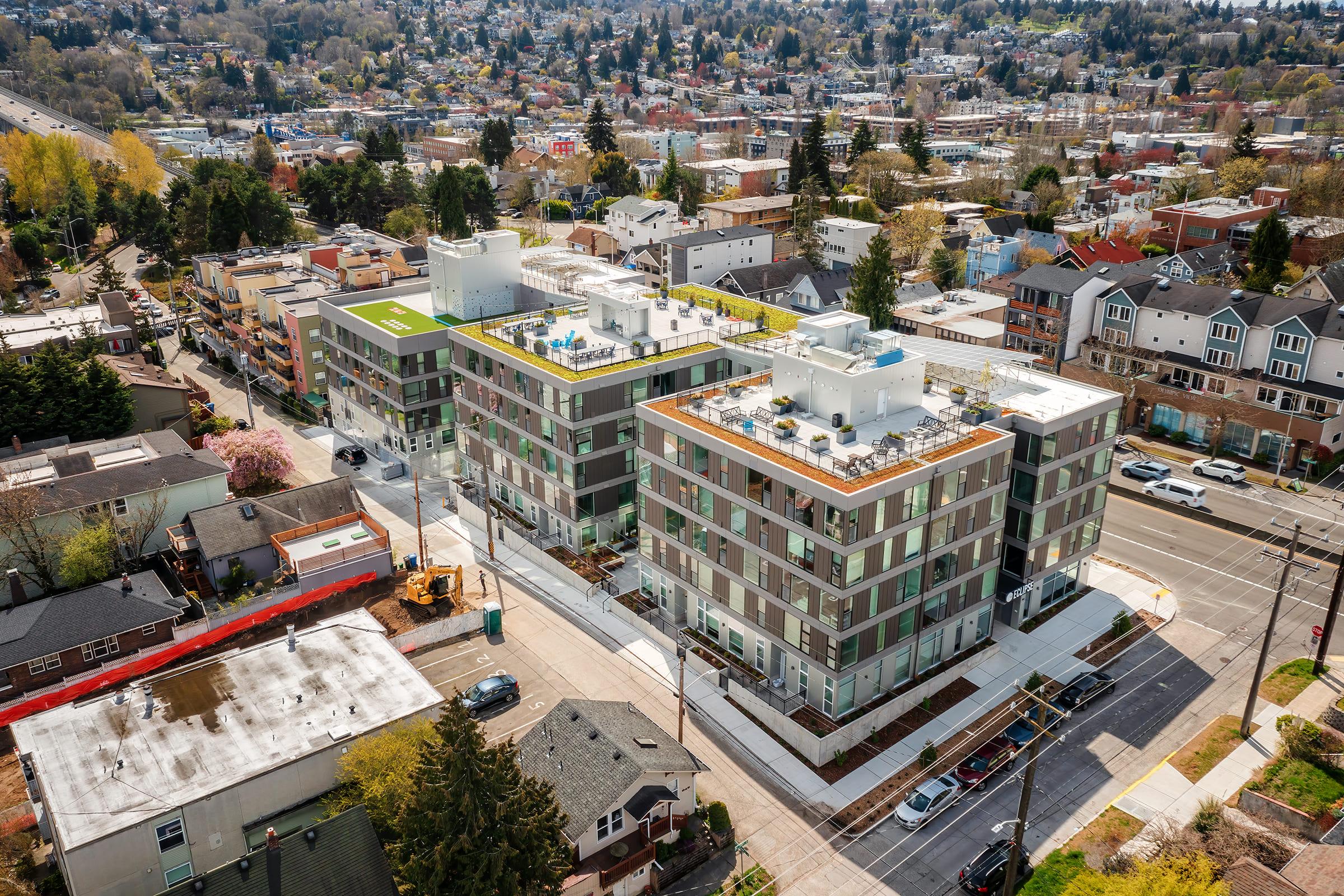 Aerial view of modern apartment buildings with green rooftops and outdoor spaces, surrounded by a mix of residential and commercial properties. The cityscape features trees and streets, showcasing urban development and landscape in a vibrant neighborhood.