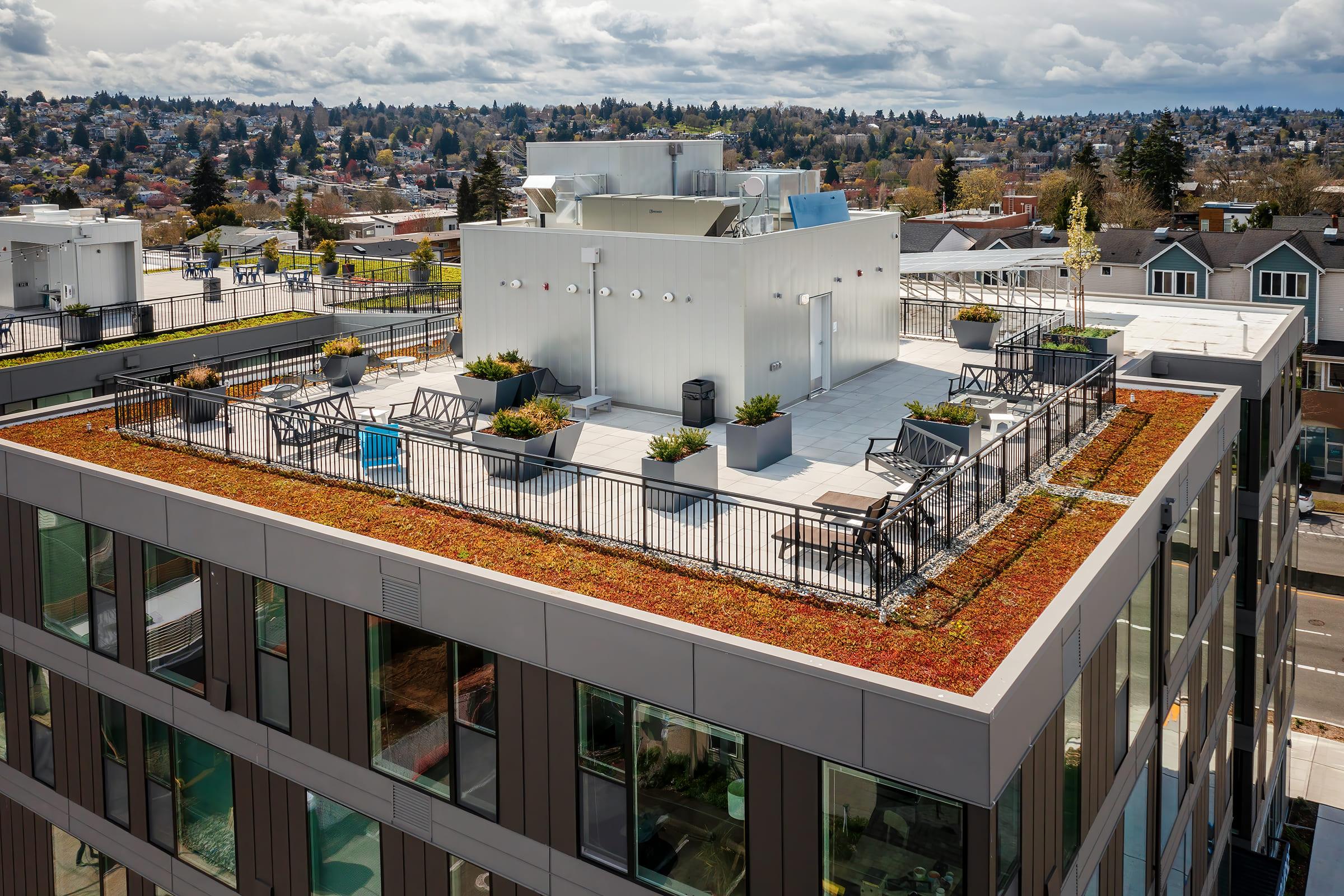 Aerial view of a modern building rooftop featuring a landscaped garden with colorful plants, several seating areas, and a white structure in the center. The surrounding skyline includes low-rise buildings and trees under a partly cloudy sky.