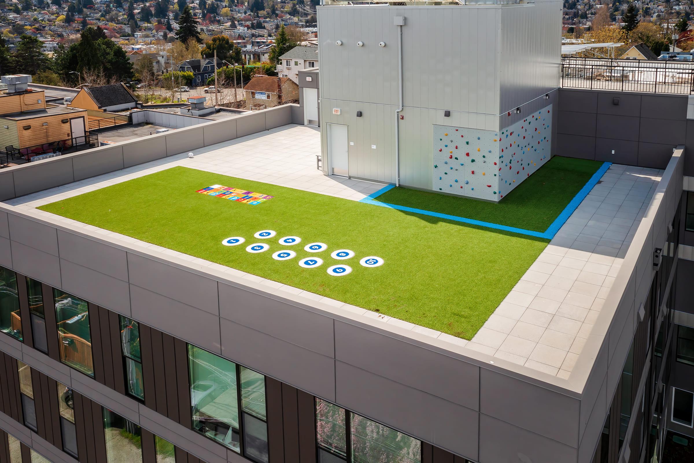 A rooftop space featuring green artificial grass, a climbing wall, and colorful game designs. The area is surrounded by a low wall, with various buildings and trees visible in the background. Natural sunlight illuminates the space, creating a vibrant recreational environment.