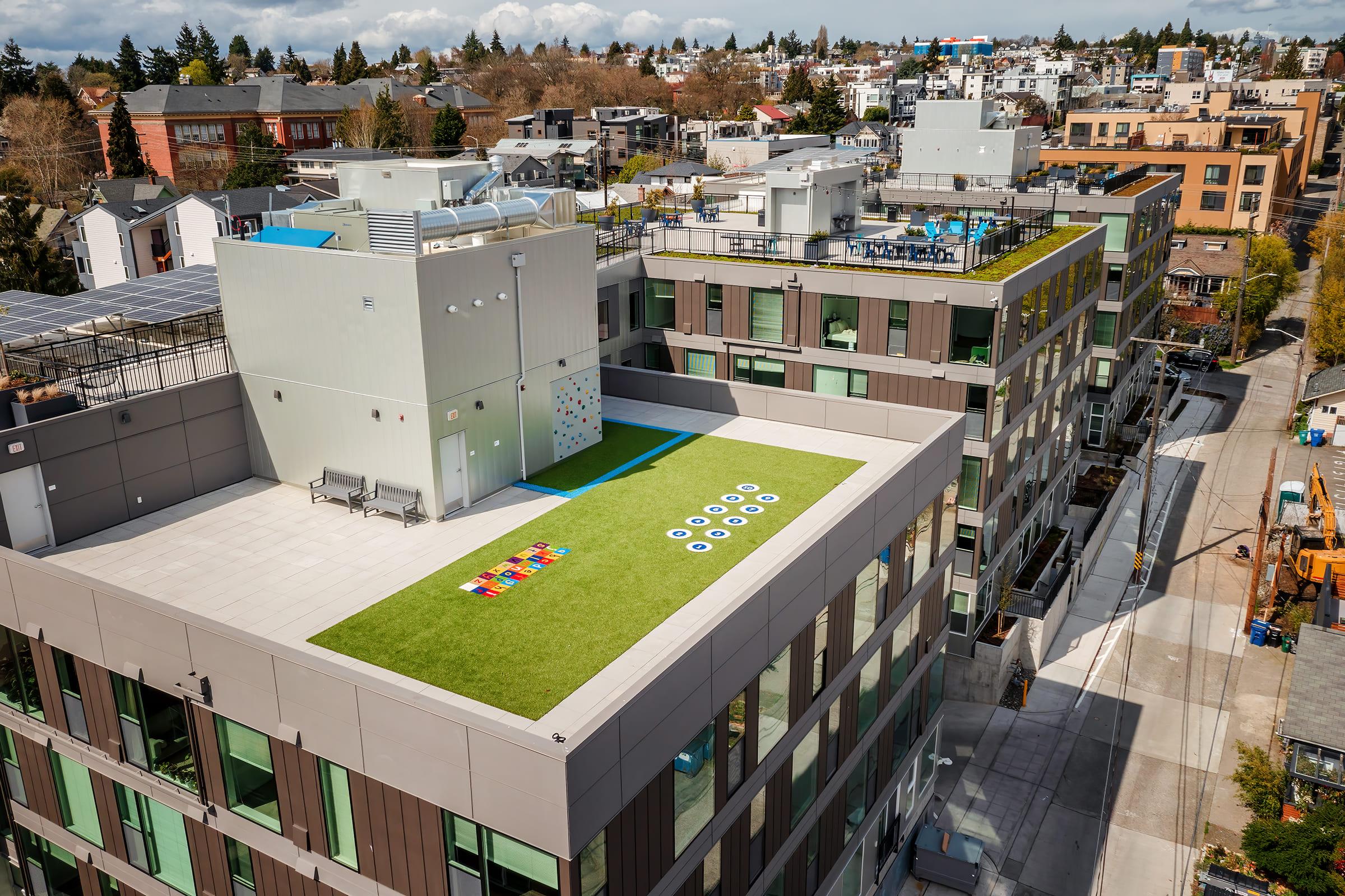 Aerial view of modern buildings with a rooftop area featuring artificial grass, play equipment, and colorful games. Solar panels are visible on the roof, and surrounding urban architecture shows a mix of residences and commercial spaces. A clear sky with clouds adds to the vibrant scene.