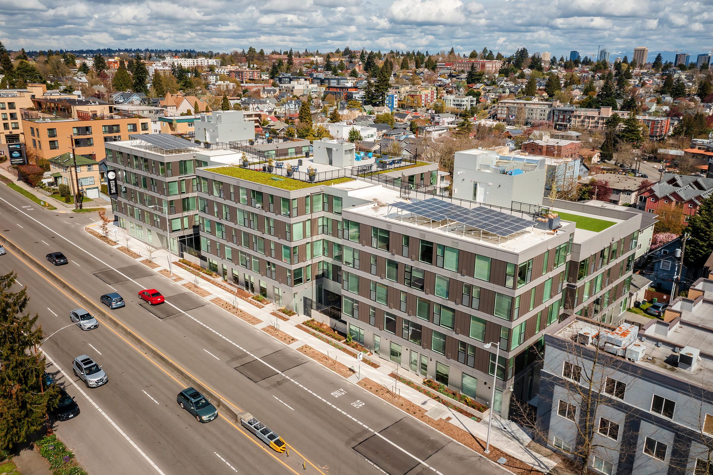 Aerial view of a modern multi-story apartment building with green roofs, alongside a busy street. The building features large glass windows and is surrounded by trees and other structures. In the background, there are multiple residential areas and a partly cloudy sky.