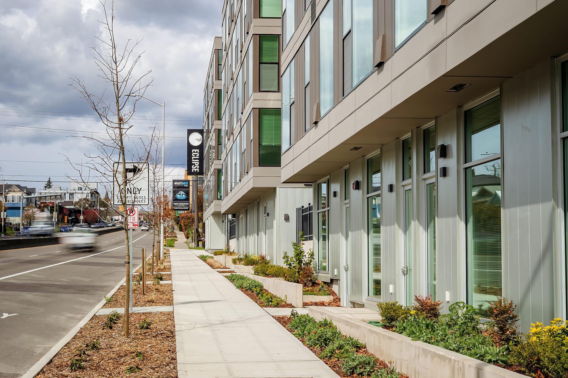 Street view of a modern building with large windows and a sign reading "Elysian." The sidewalk is lined with small plants and trees, and a road runs alongside the property where a vehicle is passing. The sky is partly cloudy, showcasing a typical urban setting.