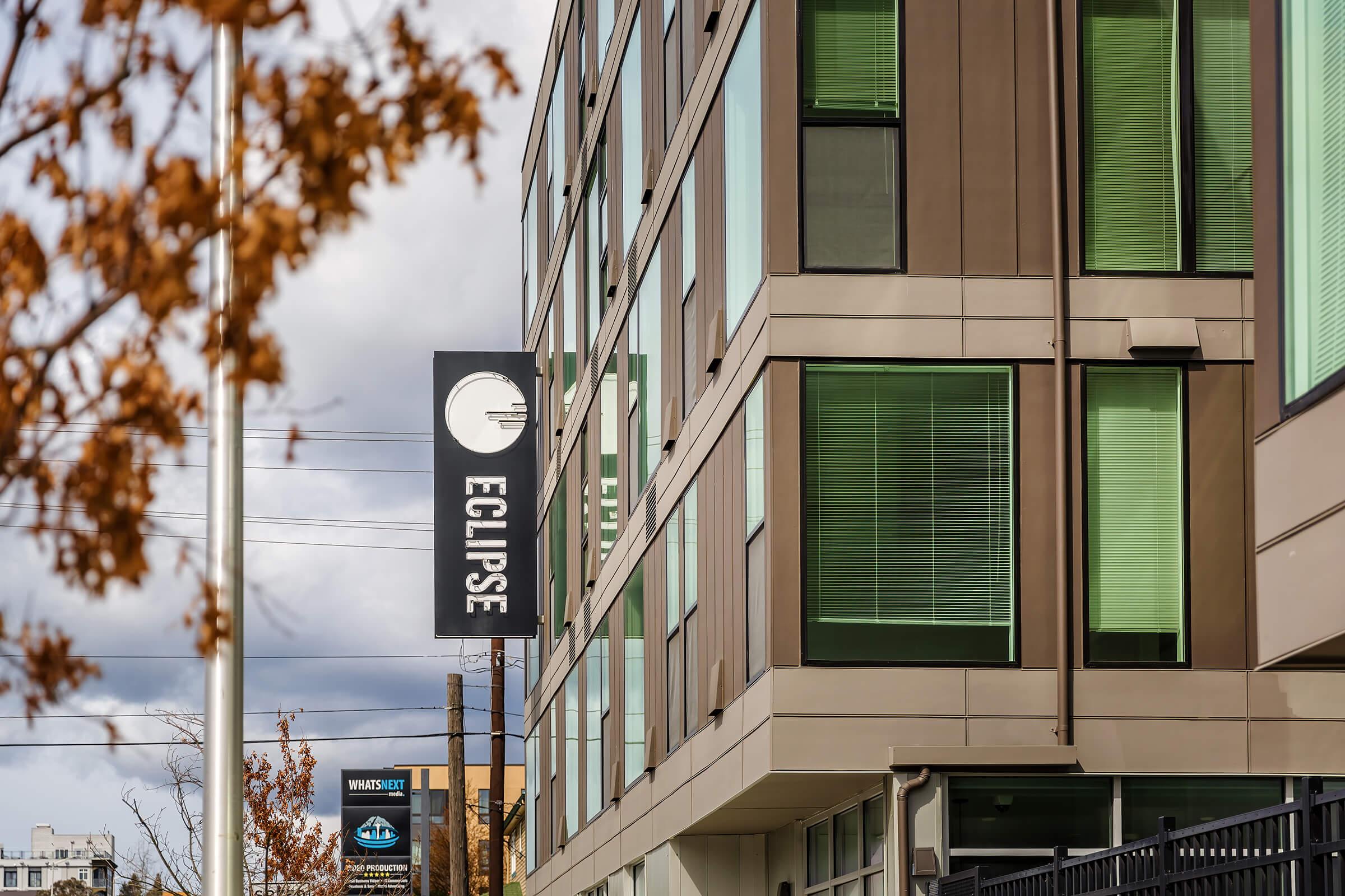 A contemporary building with large green-tinted windows and a sign that reads "ECLIPSE." The sign features a circular design. In the background, there are utility poles and additional signage partially visible. The scene is set against a cloudy sky with autumn foliage nearby.