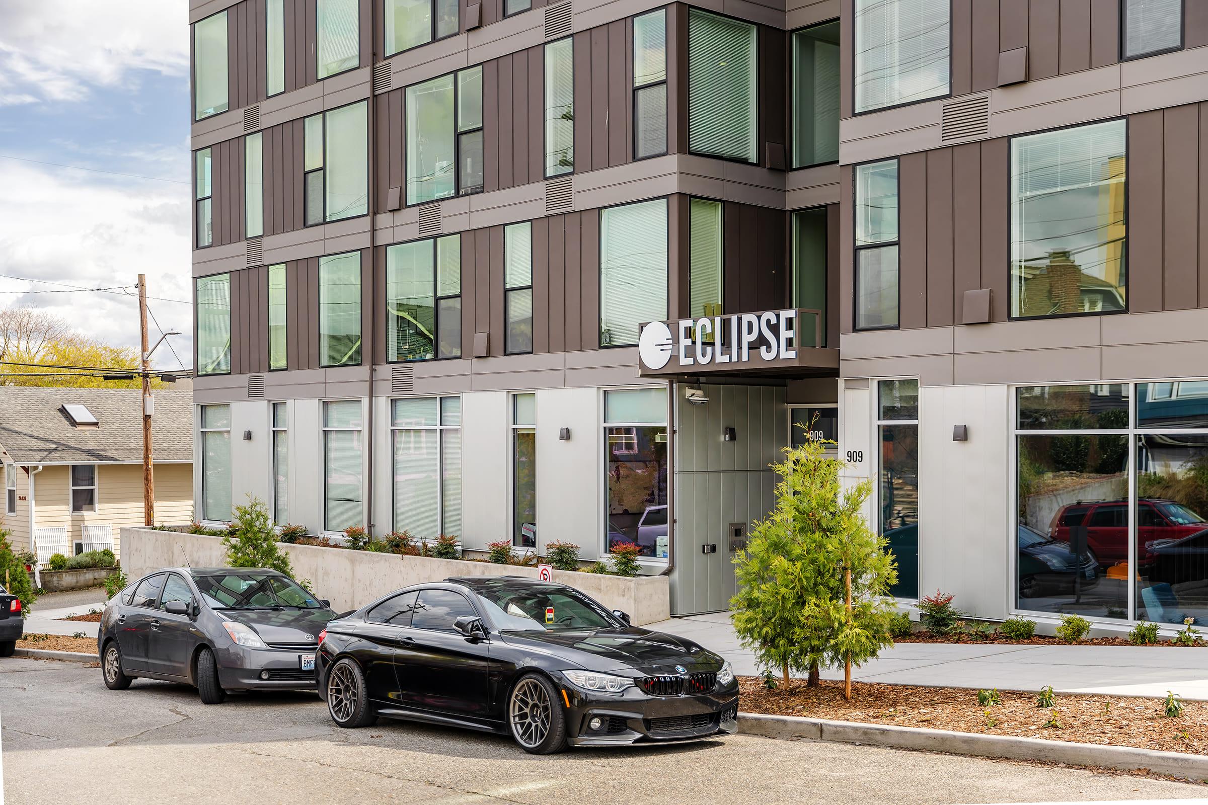 A sleek black car parked in front of a modern apartment building named "Eclipse." The building features large windows and a contemporary design, surrounded by landscaped greenery. Another car is parked nearby, and the scene is set on a sloped street under a partly cloudy sky.