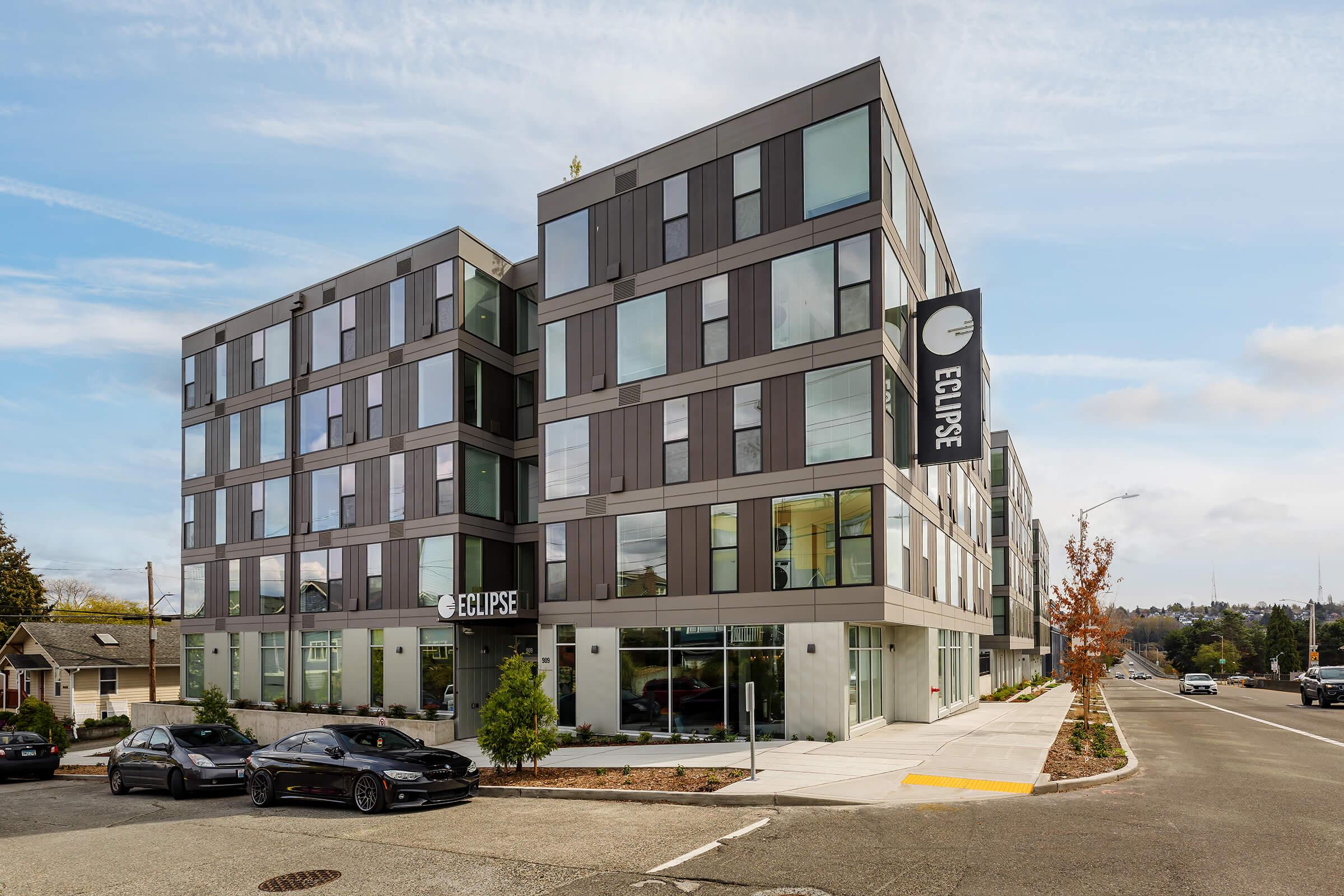 Modern multi-story building named "Eclipse," featuring large glass windows and a sleek design. The building has a black and white color scheme, and a sign displaying the name. Two black cars are parked in front, with a sidewalk and some greenery visible. The sky is partly cloudy.