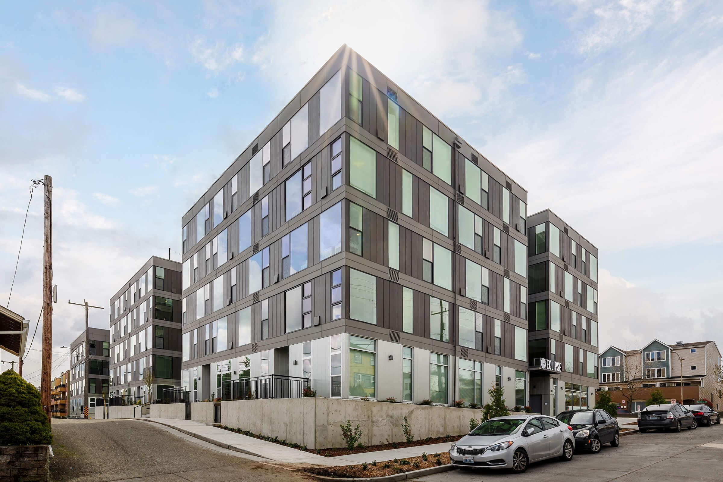 Modern multi-story residential building with a sleek design, featuring large windows and a mix of gray and green facade elements. The structure is situated on a cleanly landscaped corner lot, with parked cars in the foreground and a clear blue sky above.