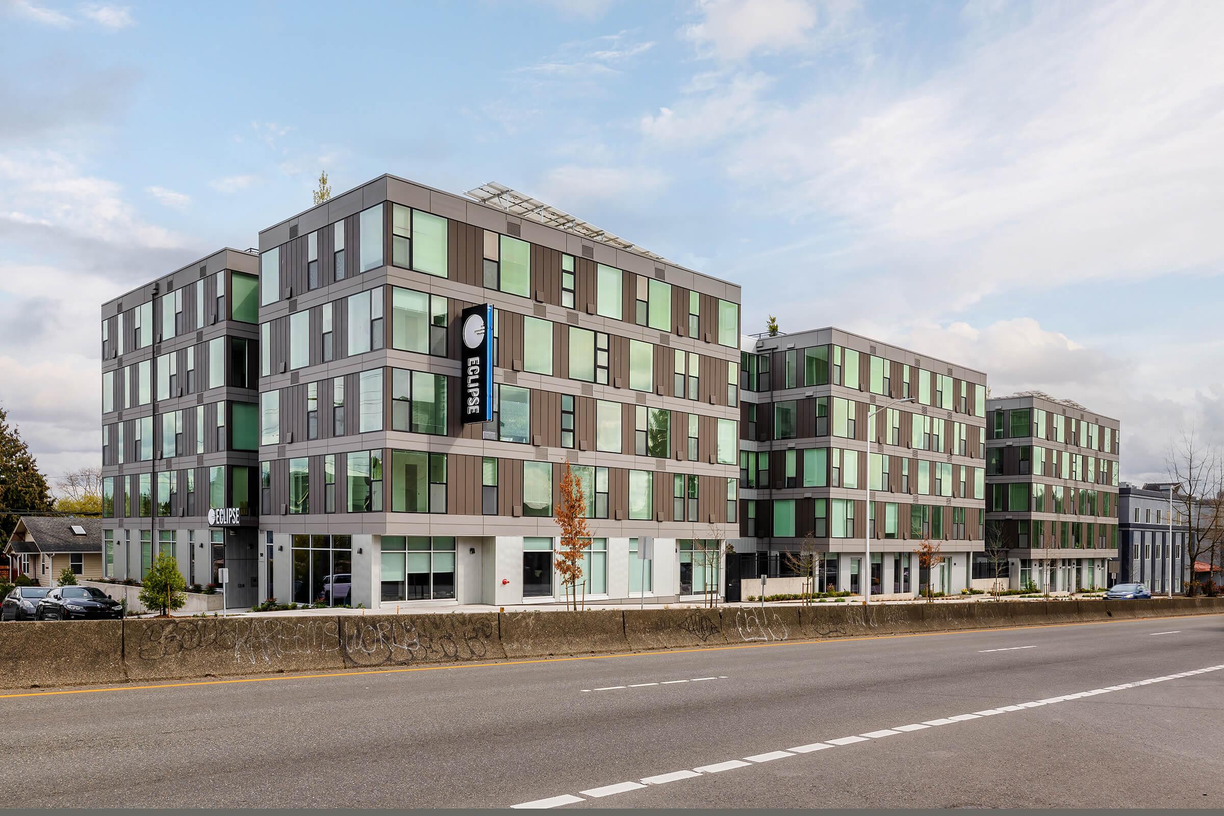 A modern apartment community featuring multiple stories with large windows and a mix of brown and gray paneling. The building has a sleek design with a sign visible in front, indicating it may be a residential or rental property. The setting includes a roadway in the foreground and trees nearby.