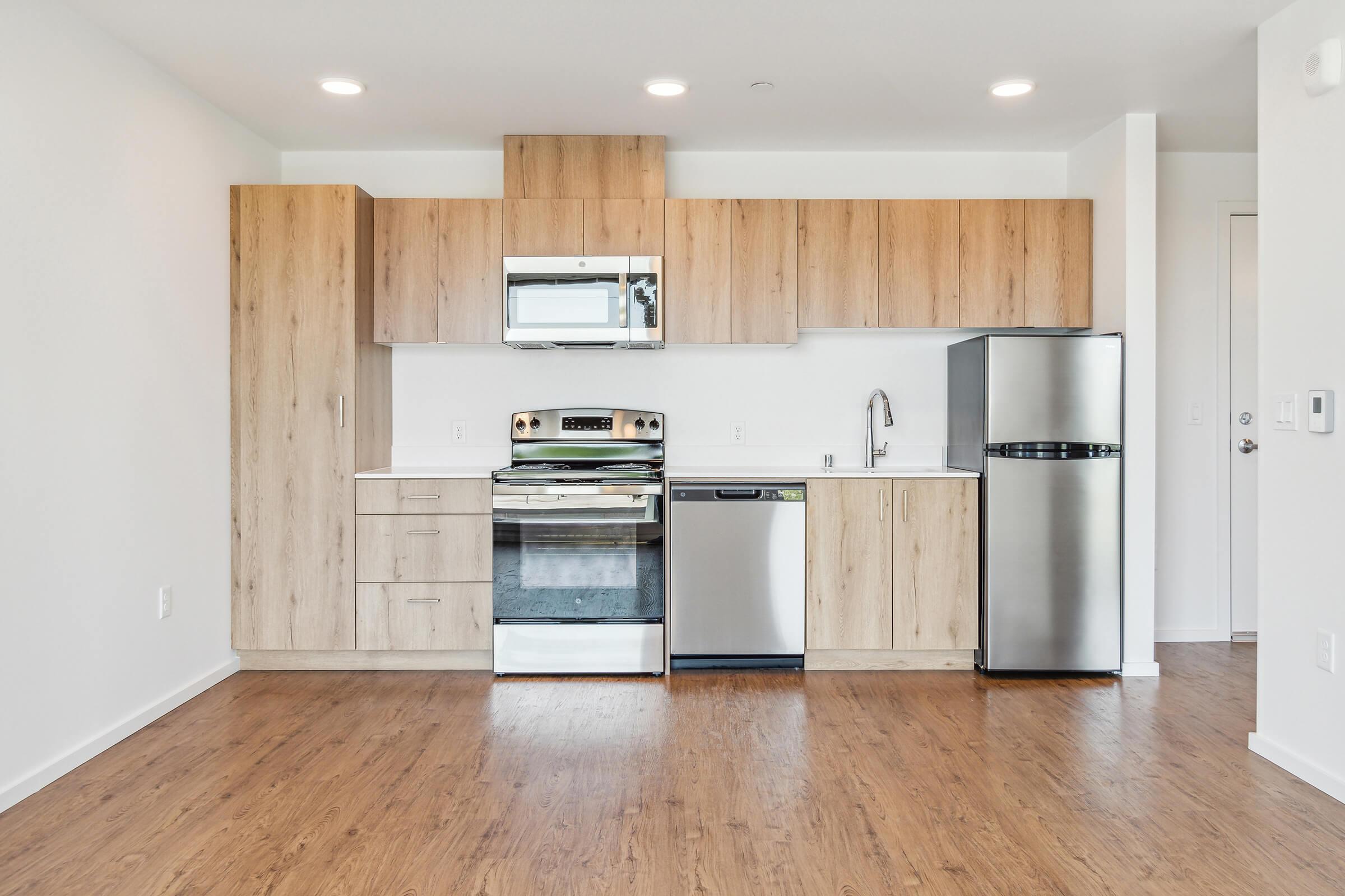 Modern kitchen featuring wooden cabinetry, stainless steel appliances including an oven, microwave, refrigerator, and dishwasher. The space has a light, open feel with wood flooring and recessed lighting, set against a bright, white wall.