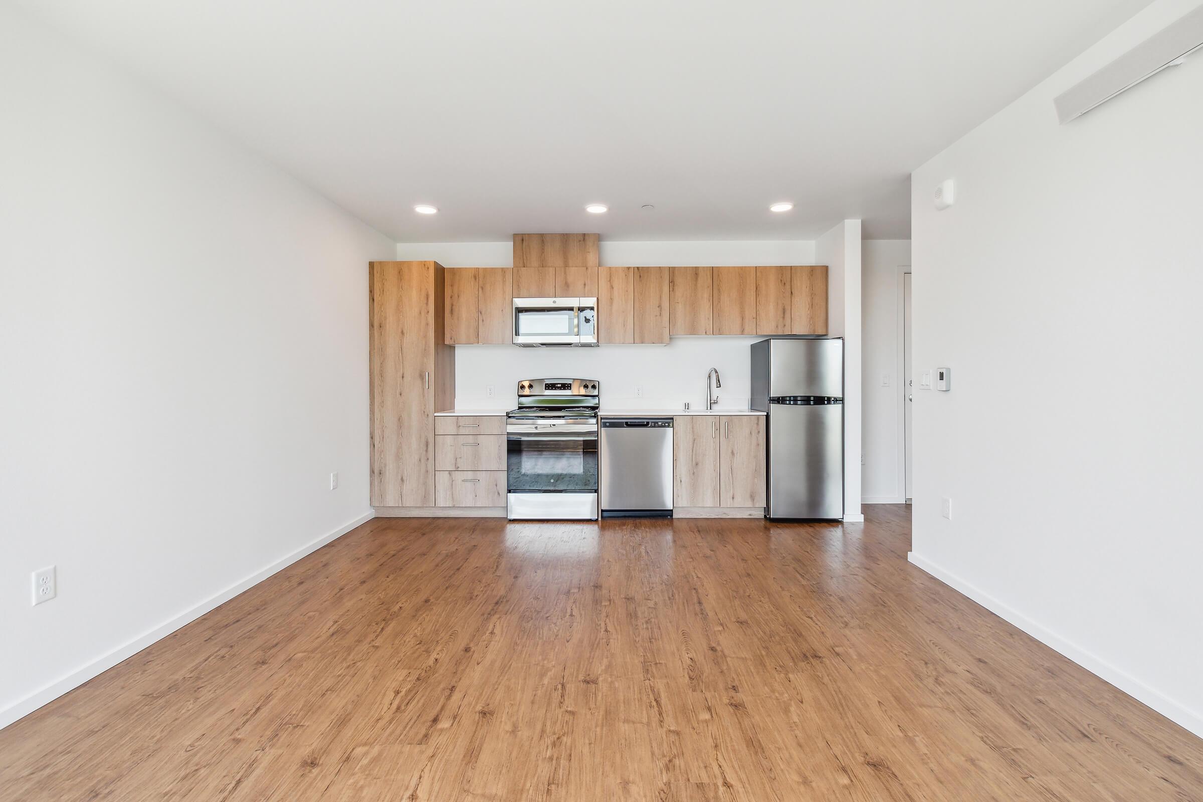 A modern kitchen area featuring light wood cabinetry, stainless steel appliances including an oven and fridge, and a spacious layout. The floor is covered in light wood laminate, and the walls are painted white, creating a bright and airy atmosphere. Natural light is present from the windows.