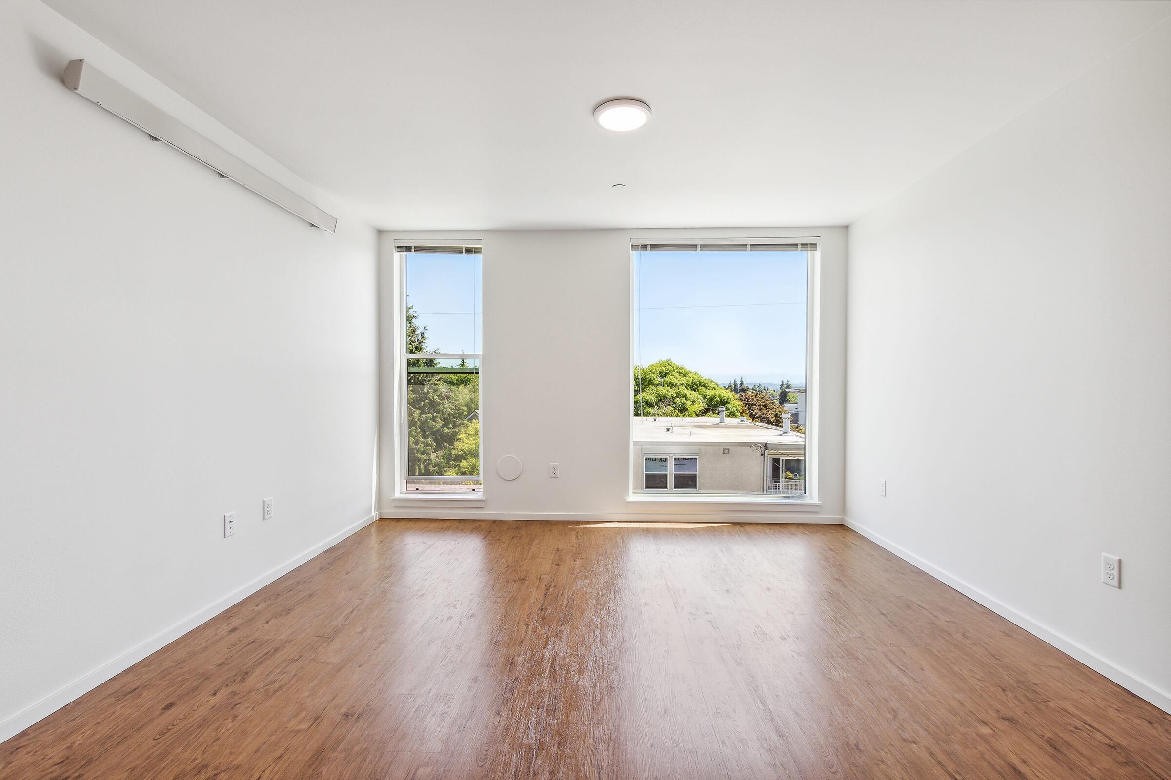 A bright, empty room with white walls and large windows. The polished wooden floor reflects natural light. One wall features an air conditioning unit. Outside the windows, green trees and a building can be seen, indicating a serene view.