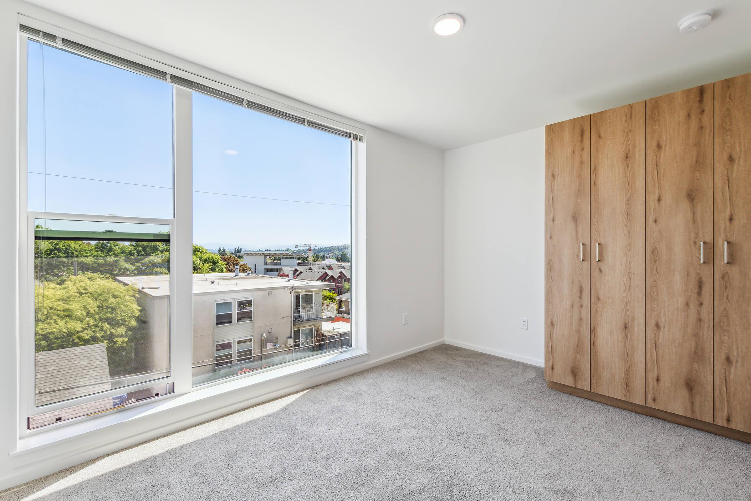 Bright, empty room with light-colored walls and a plush carpet. A large window offers a view of trees and distant buildings, allowing natural light to flood the space. A wooden wardrobe stands against one wall, adding a touch of warmth to the modern, minimalist design.