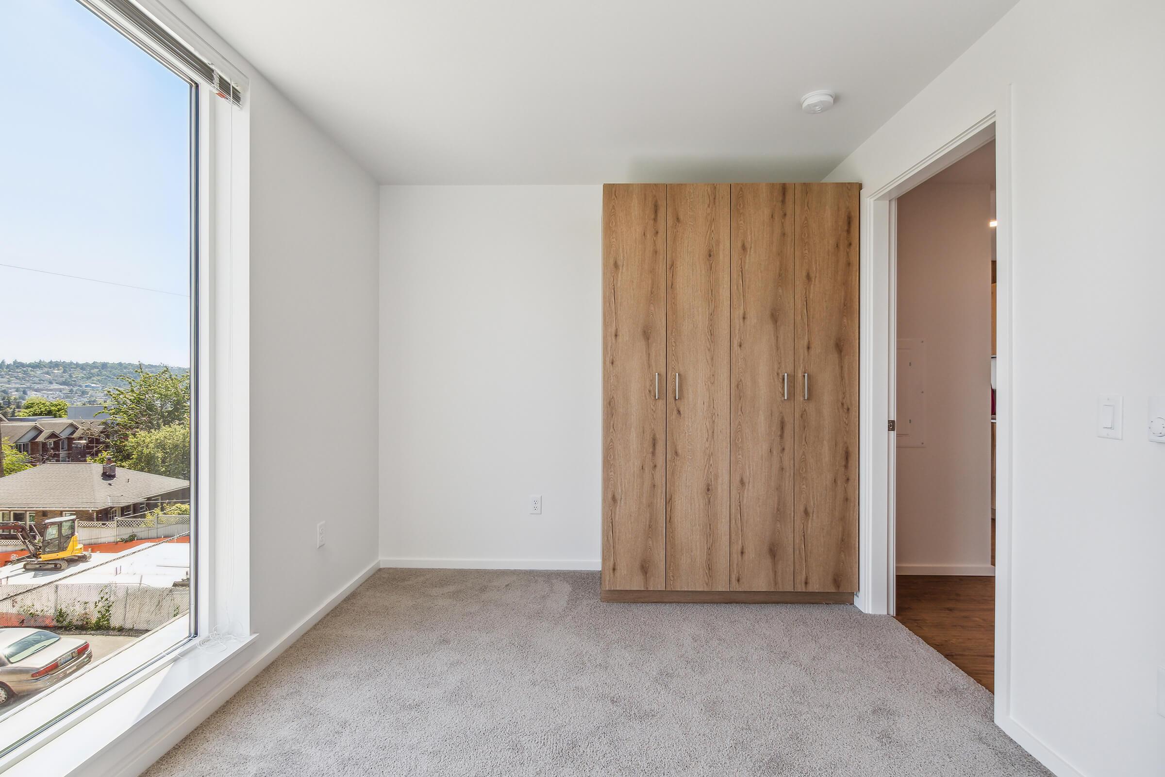 Interior view of a small, modern room featuring a large window with a scenic view, soft beige carpet flooring, and a wooden wardrobe against one wall. The walls are painted in a light color, creating a bright and airy atmosphere. There is a doorway leading to another room.