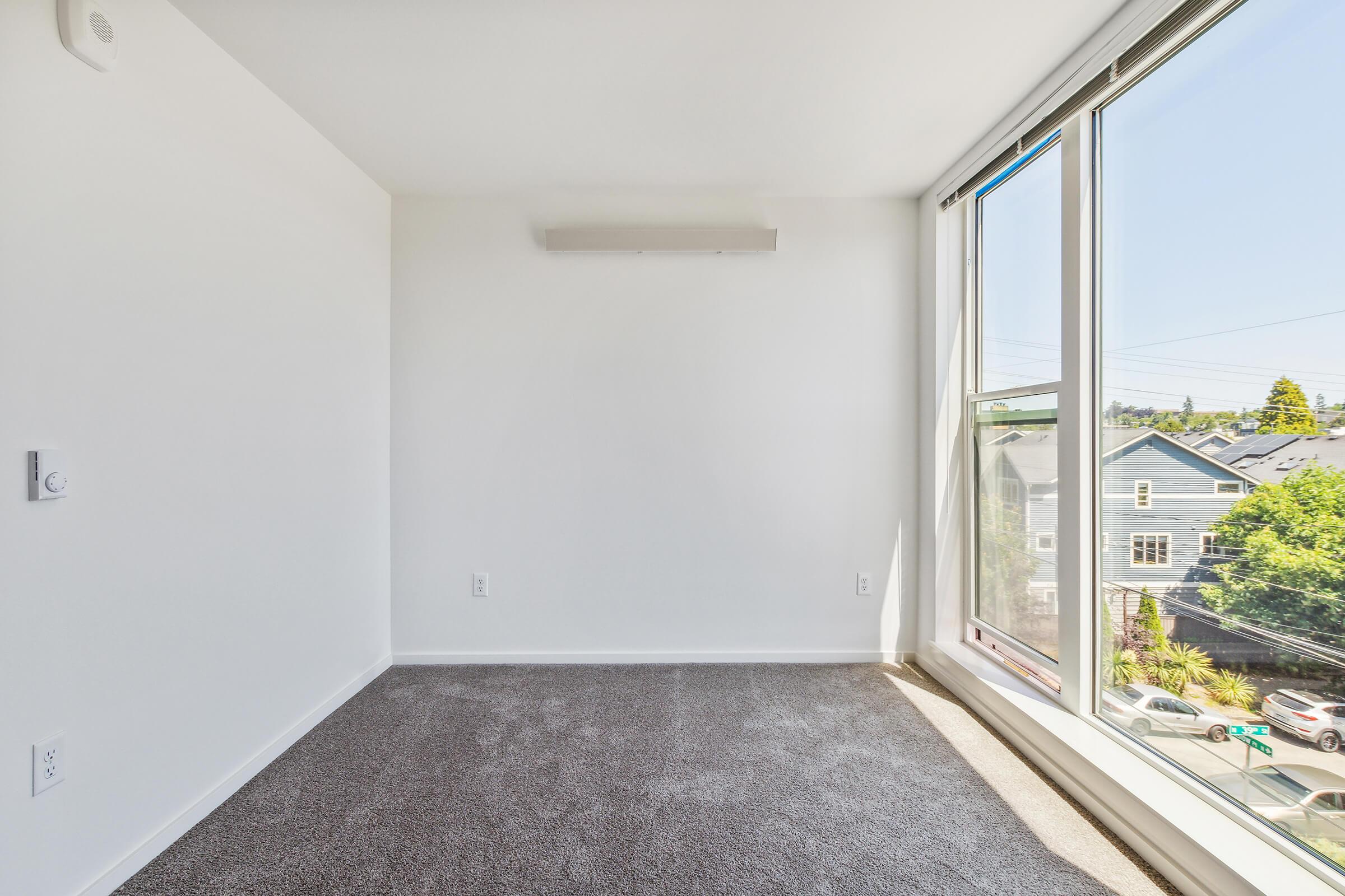 A minimalist room with light-colored walls and soft gray carpet. A large window lets in natural light and offers a view of neighboring houses. The room features no furniture, highlighting its spaciousness and potential for customization.