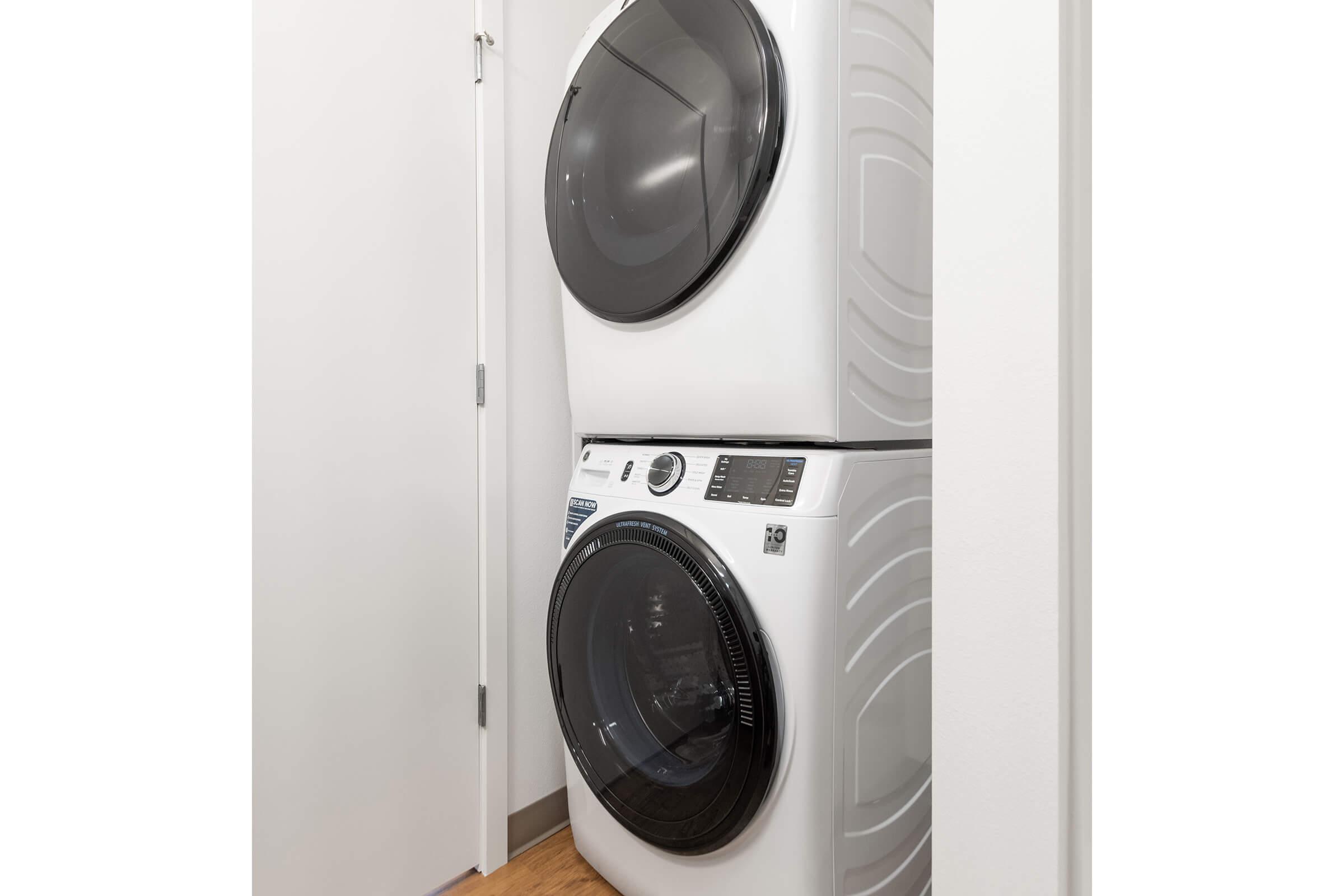 Stacked white washer and dryer set in a narrow laundry space, with a closed door nearby. The appliances have a modern design, featuring a digital control panel on the front of the washer. The floor is made of light wood, complementing the minimalist aesthetic of the room.