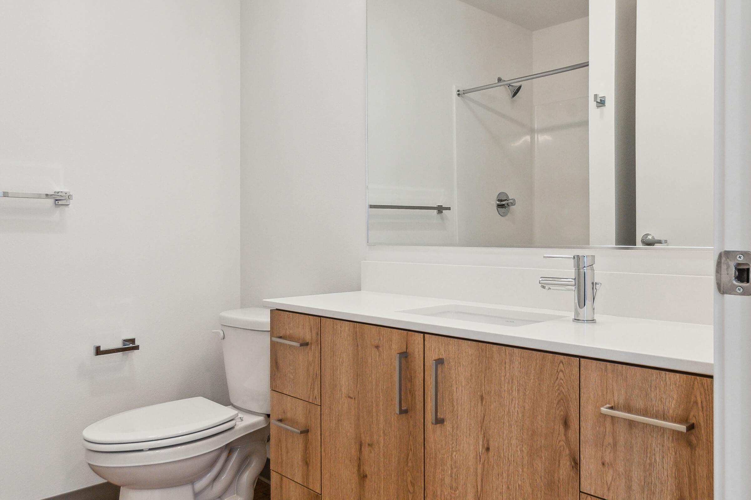 Modern bathroom featuring a white countertop and wooden cabinetry. A sleek faucet sits above a minimalist sink. To the left, there is a toilet, and a large mirror reflects the space. A glass shower enclosure is visible in the background, adding to the contemporary design.