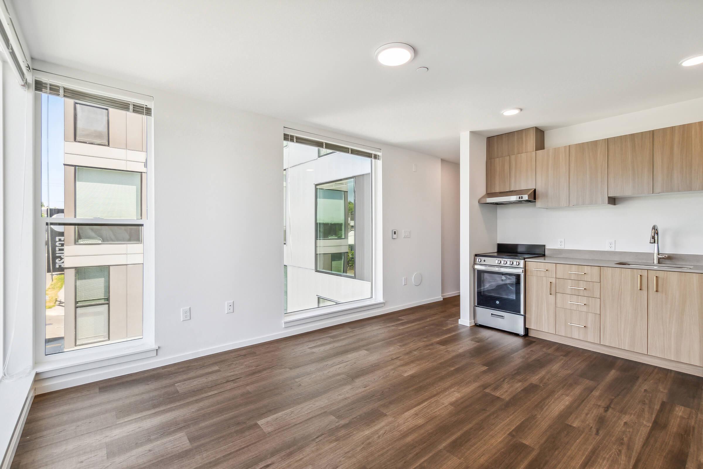 Modern, bright apartment featuring a spacious living area with natural light, large windows, and a sleek kitchen. The kitchen includes wood cabinets, a stove, and ample counter space. The flooring is light-colored wood, creating a warm atmosphere in the open layout.