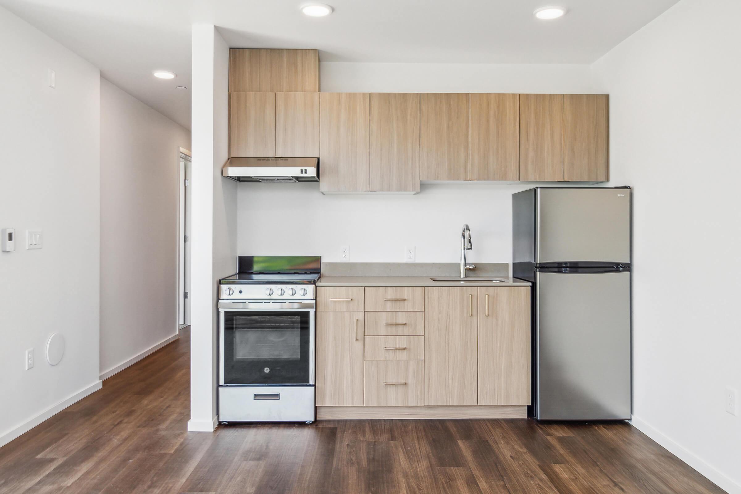 A modern kitchenette featuring a stove, oven, and range hood. The cabinetry above is light wood, while the lower cabinets have a minimalist design. A sleek refrigerator stands beside the counter, and the flooring is a warm wood tone. The walls are painted white, creating a bright and spacious feel.