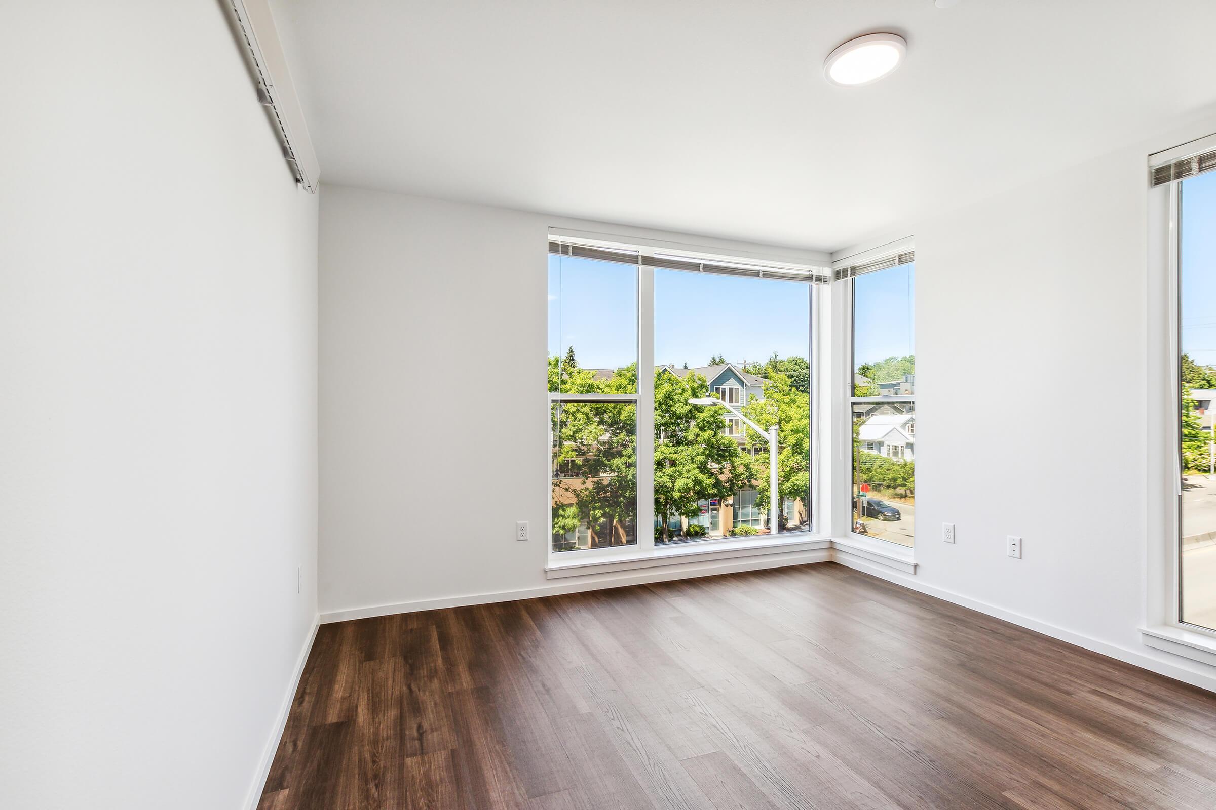 A bright, empty room with white walls and large windows allowing natural light to flow in. The floor is made of dark wood, and there are no furnishings. Outside the windows, green trees and residential buildings are visible, creating a peaceful urban view.