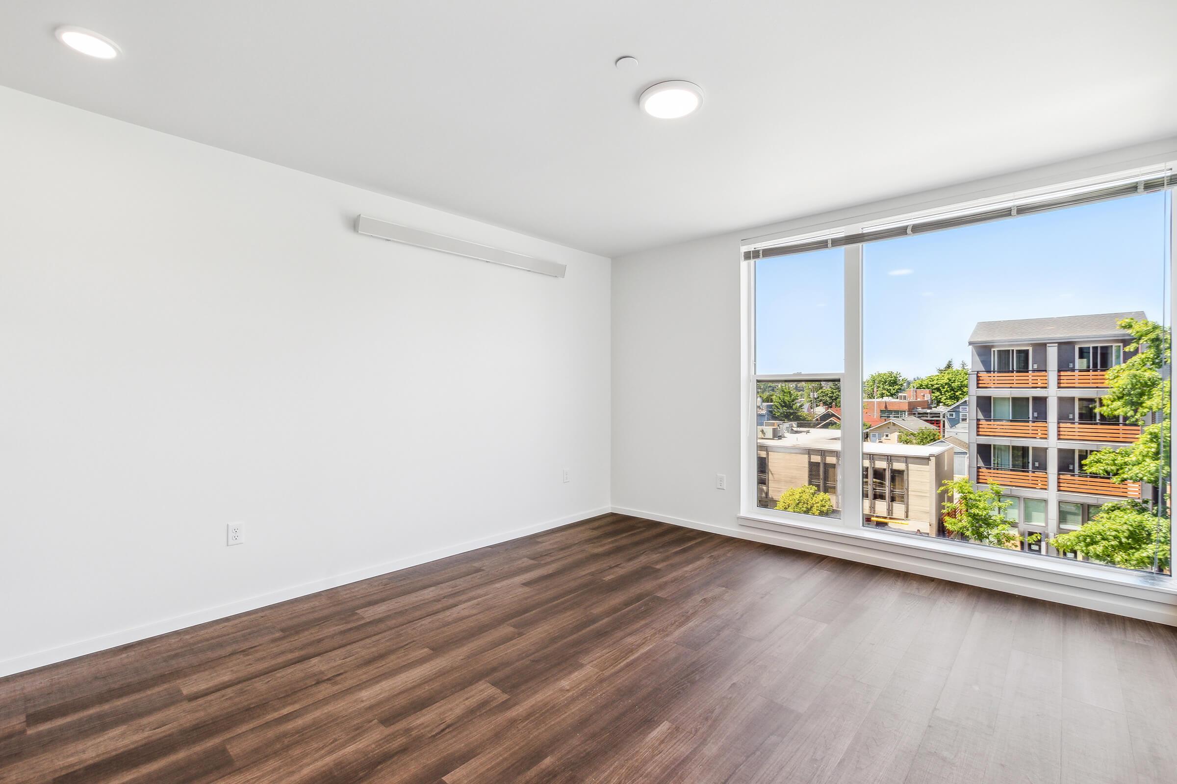 A spacious, empty room with light-colored walls and hardwood floors. A large window provides natural light and a view of neighboring buildings and greenery outside. The ceiling features modern light fixtures, and the overall atmosphere is bright and airy.