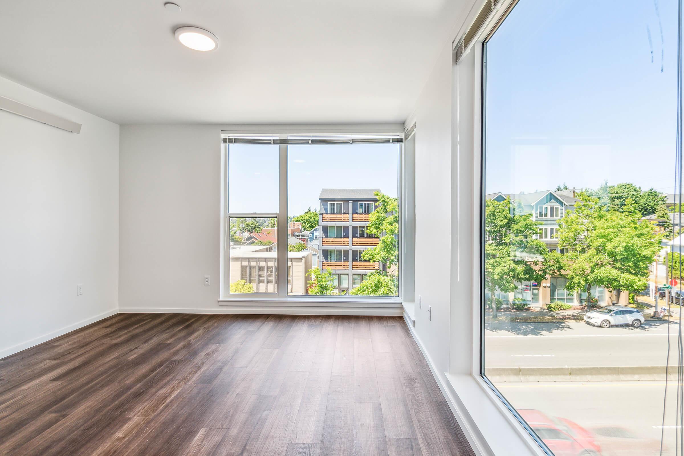 Bright, empty room with large windows showcasing a view of trees and buildings outside. The space features hardwood flooring and white walls, providing a clean and modern aesthetic. Natural light fills the room, creating an inviting atmosphere. 