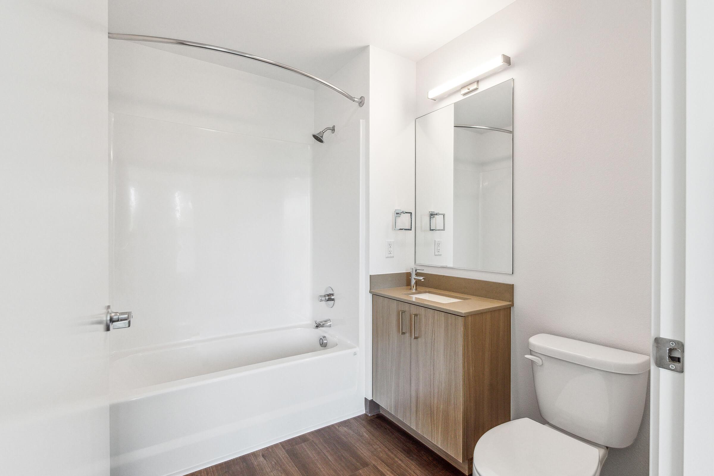 A modern bathroom featuring a white bathtub with a shower curtain, a wooden vanity with a sink and mirror, and a toilet. The walls are light-colored, and the floor has dark wood-like tiles, creating a clean and minimalist aesthetic. Natural light comes from a nearby window.