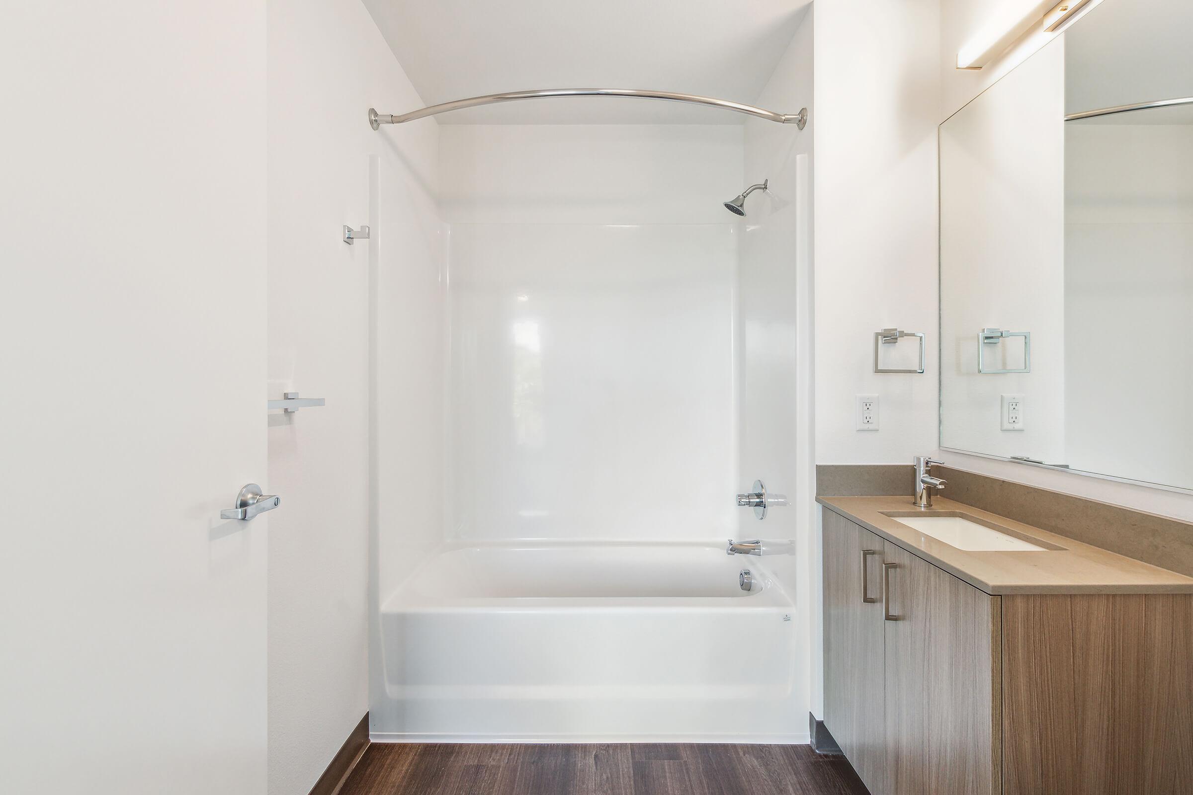 Modern bathroom featuring a bathtub with a curved shower rod, a single wall-mounted faucet, and a large mirror above a wooden vanity. The walls are painted white, and the floor is covered with dark wood. Soft natural light brightens the space.