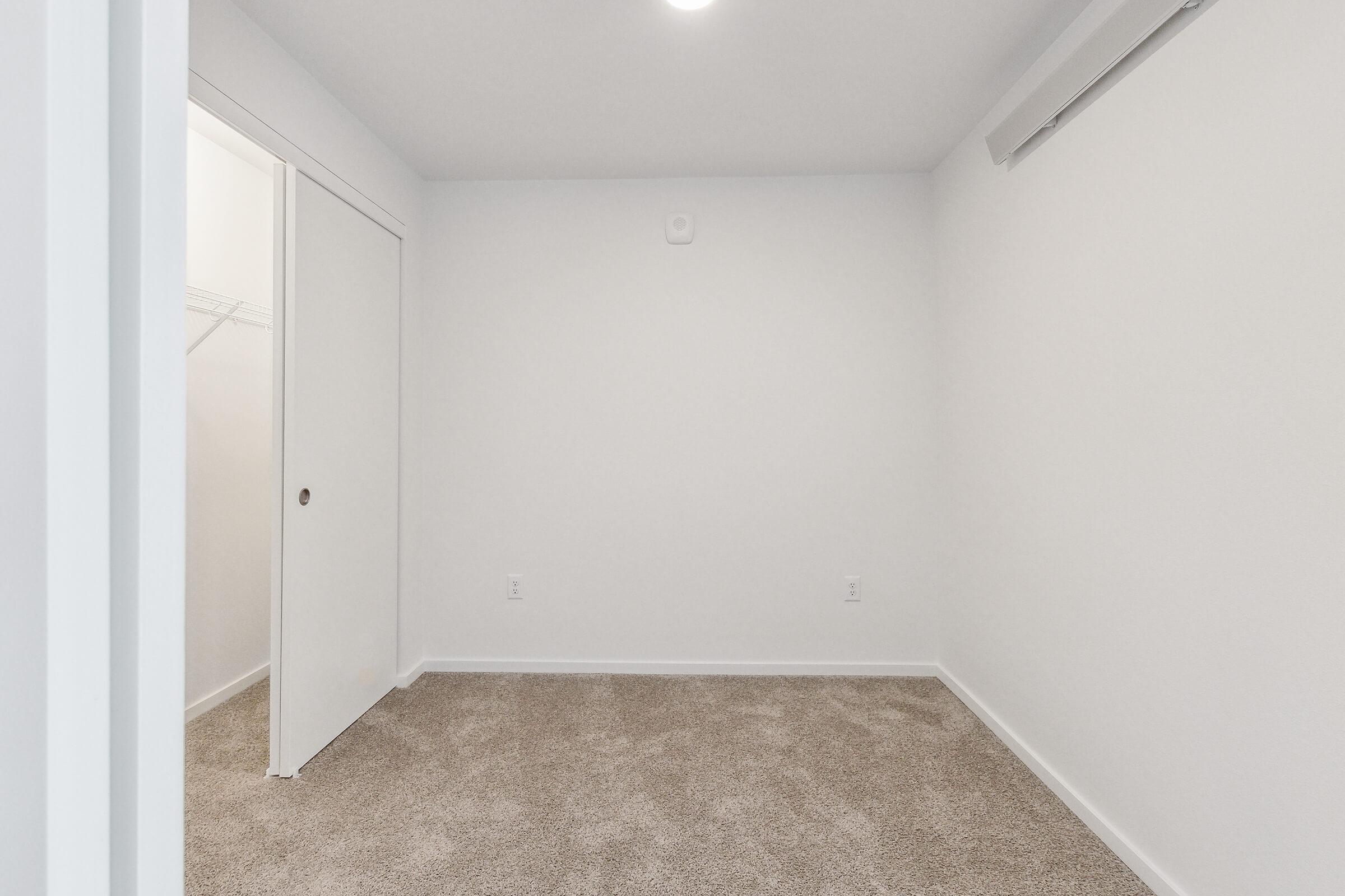 A spacious, empty room featuring light-colored walls and beige carpet. A single white door leads to a closet on the left, and there’s an air vent on the wall. The ceiling is bright with a ceiling light, creating a clean and modern look. The room is well-lit and minimalist, ideal for various uses.