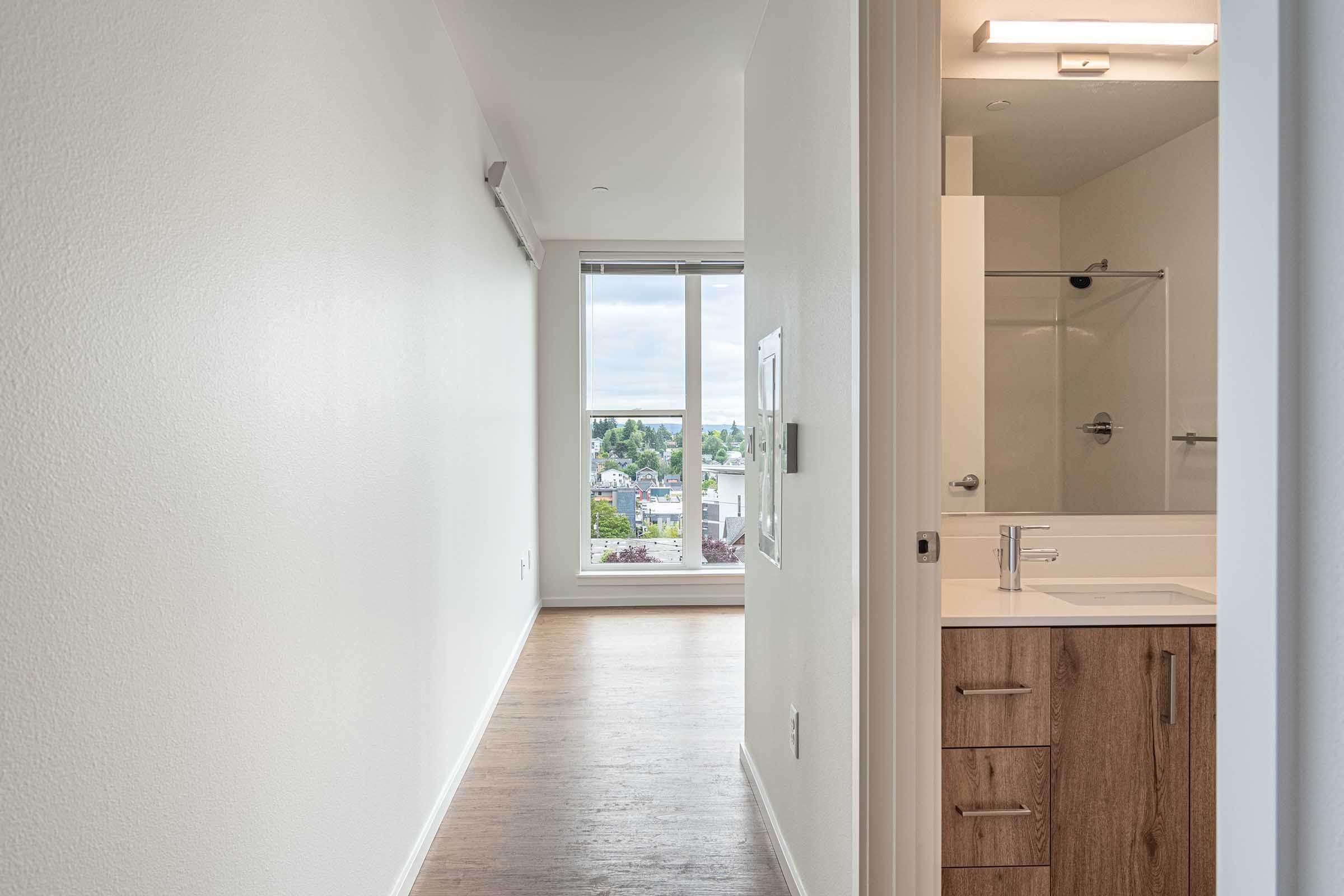 A view down a modern hallway leading to a bright, open space with large windows overlooking a cityscape. The hallway has neutral-colored walls and wood flooring, while a bathroom area with a sink and cabinet is visible on the right. Natural light fills the scene, creating a welcoming atmosphere.