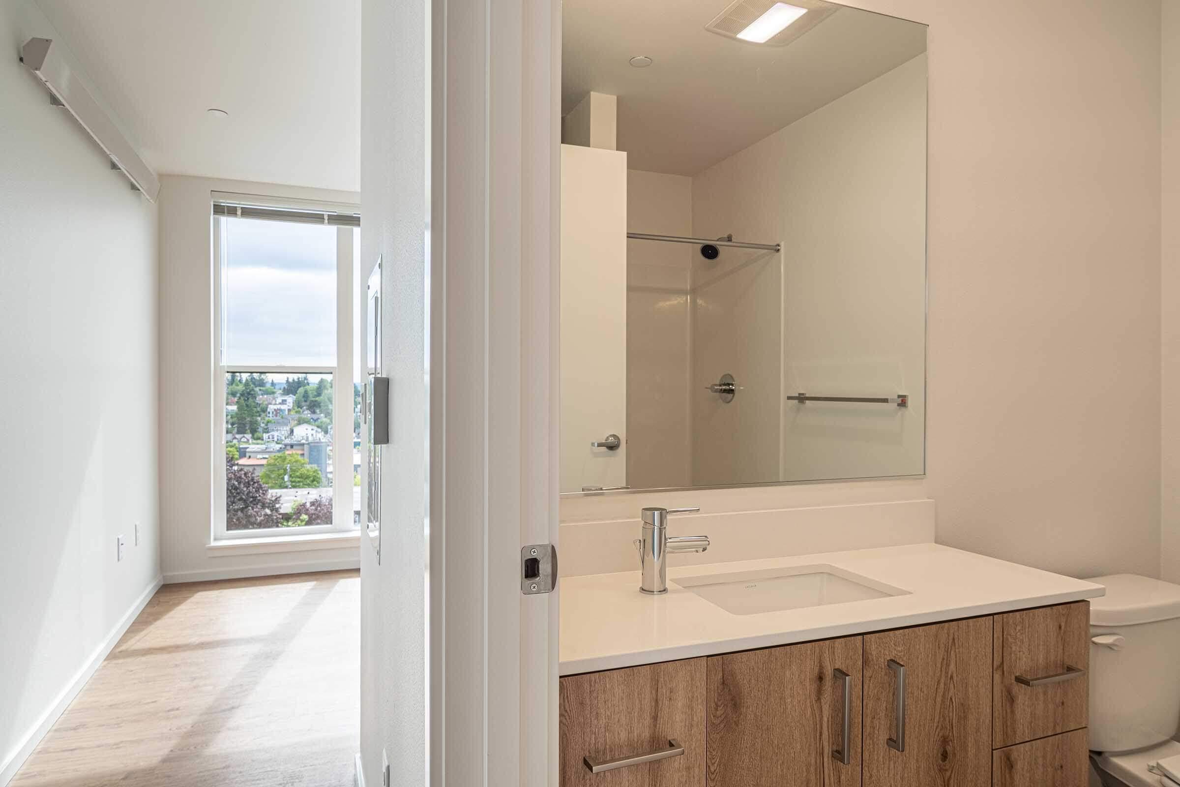 Modern bathroom with a white countertop and wooden cabinetry, featuring a mirror and a glass shower enclosure. In the background, a well-lit room with large windows showcasing a view of the outdoors. The space has light-colored walls and wooden flooring, creating a bright and airy atmosphere.