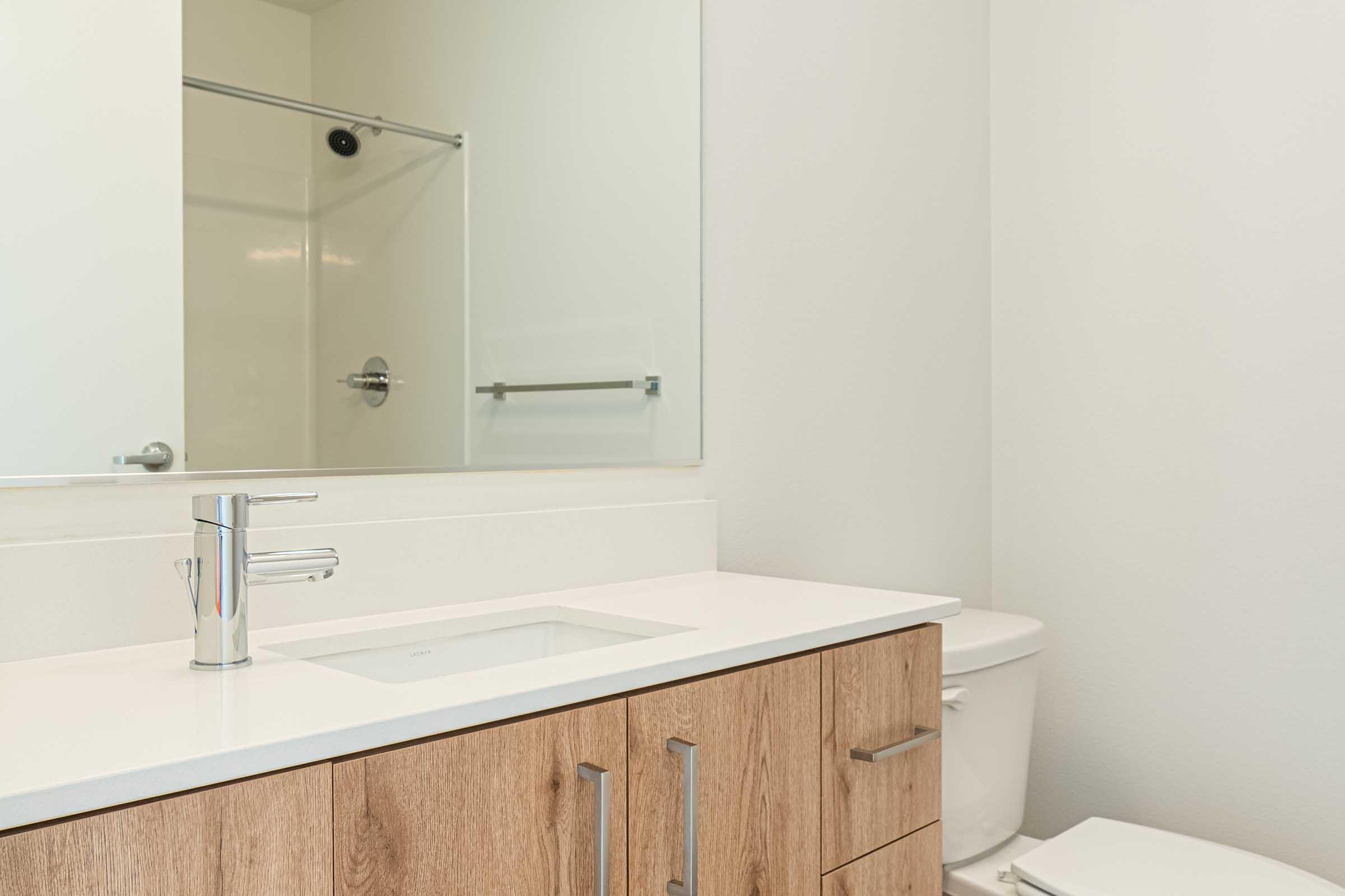 A modern bathroom featuring a sleek vanity with wooden cabinetry, a white countertop, a lavabo with a chrome faucet, a mirror above the sink, and a toilet. There is a glass shower enclosure visible in the background, set against light-colored walls.