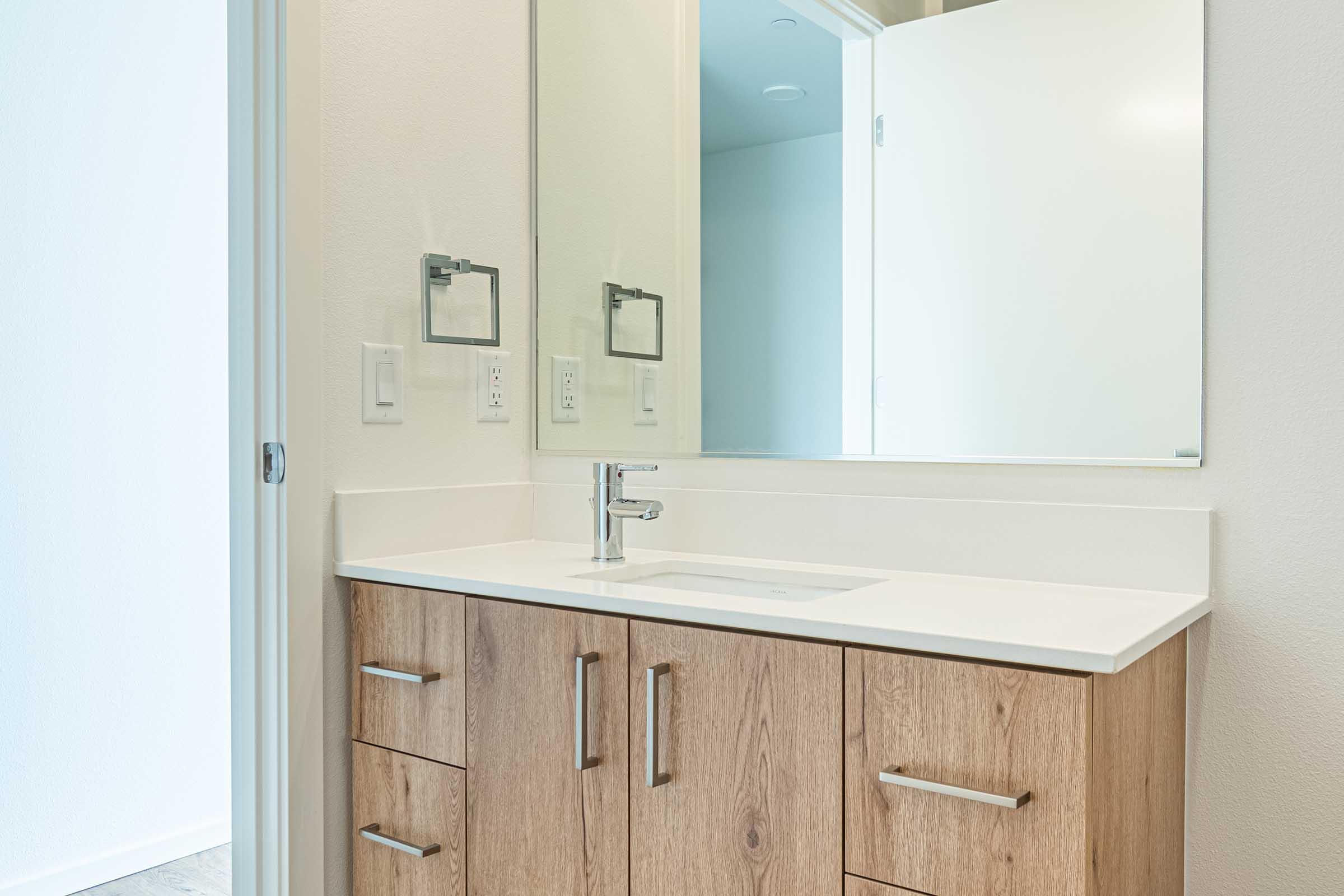 A modern bathroom featuring a minimalist design with a wooden vanity, a white countertop, and a sink. A large mirror hangs above the sink, reflecting the space. The walls are light-colored, and a door is visible on the left leading to another area. The overall aesthetic is clean and contemporary.