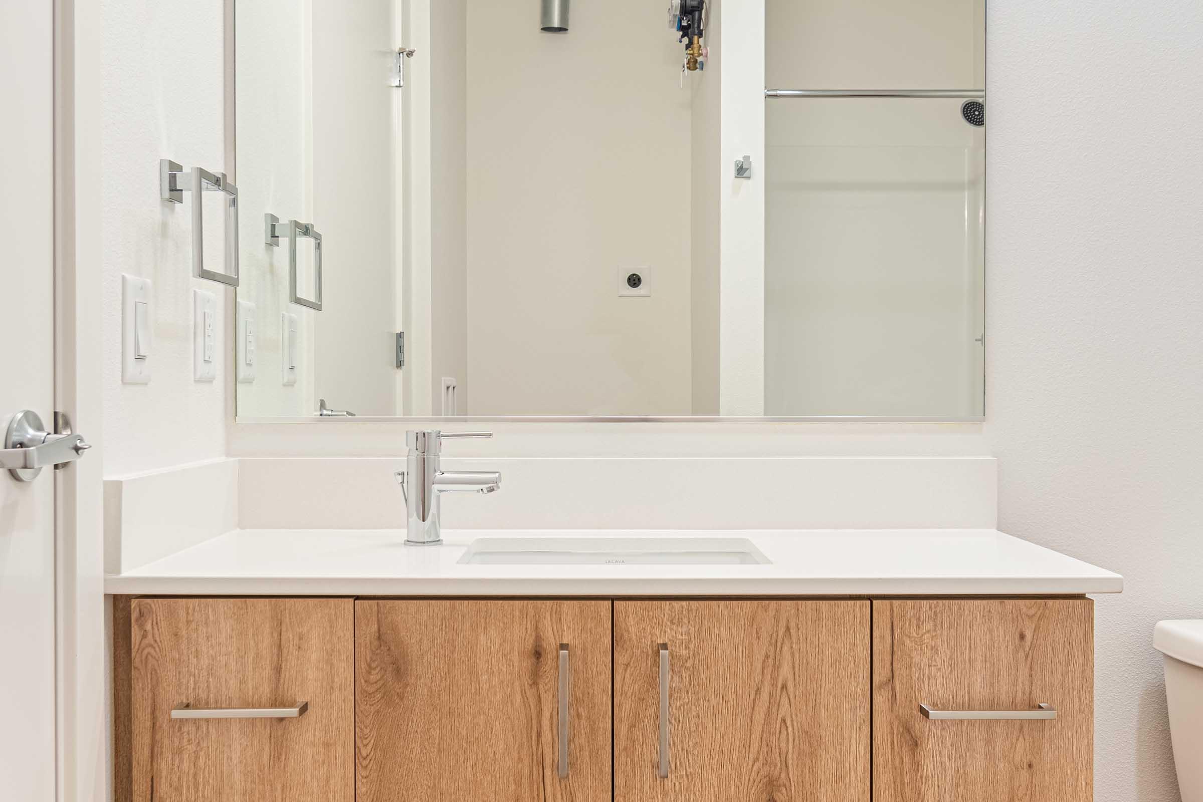 Modern bathroom vanity featuring a white countertop and a rectangular sink. Below the countertop, there are wooden cabinet doors with a natural finish. A large mirror hangs above the sink, reflecting the light and the room. The walls are painted in a light shade, creating a clean and spacious atmosphere.