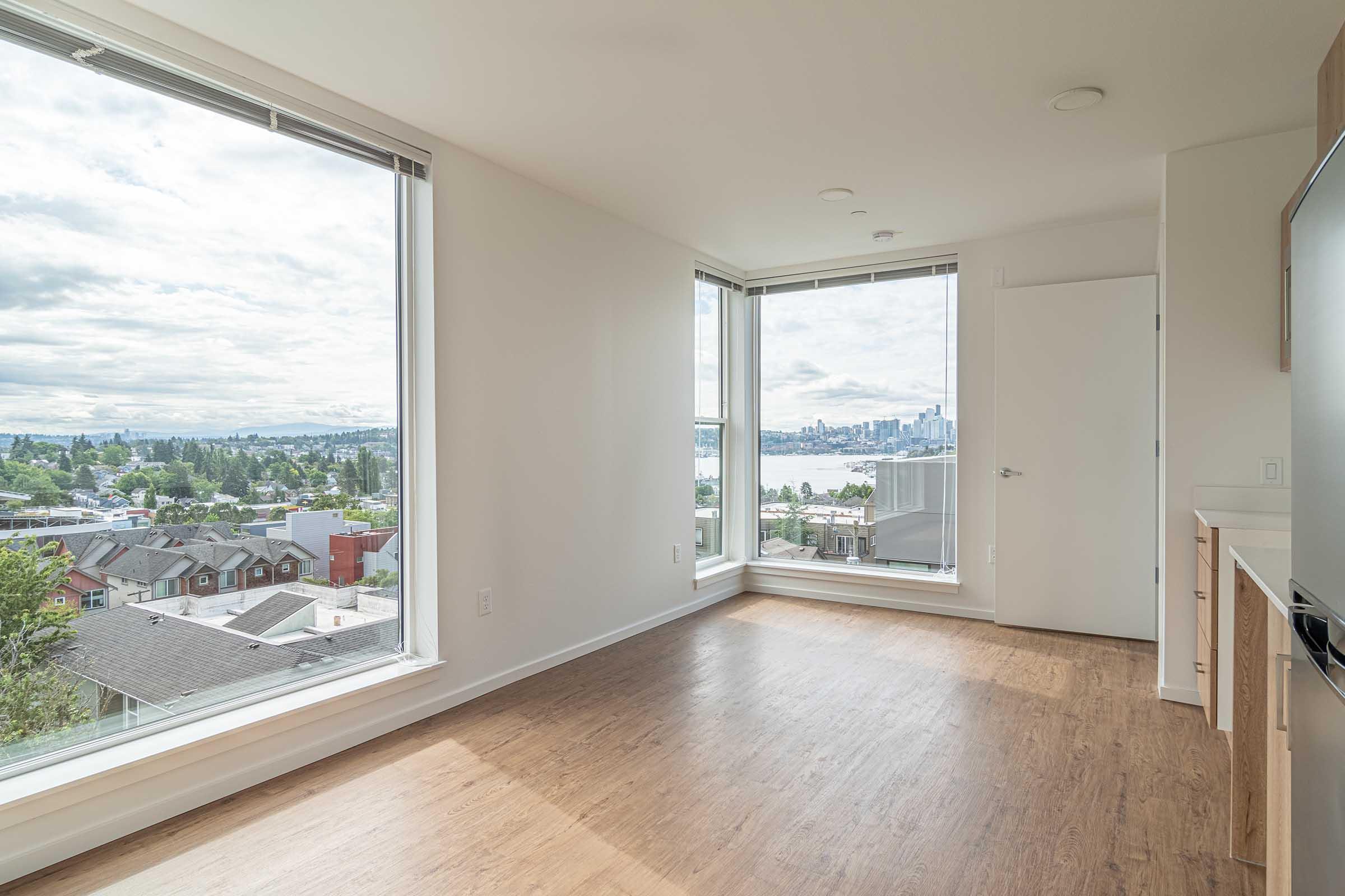 Bright, empty room with large windows offering a panoramic view of a city skyline and water. The walls are painted white, and the floor is light wood. A small kitchenette is visible on the left. The scene is well-lit by natural light, creating an open and airy atmosphere.