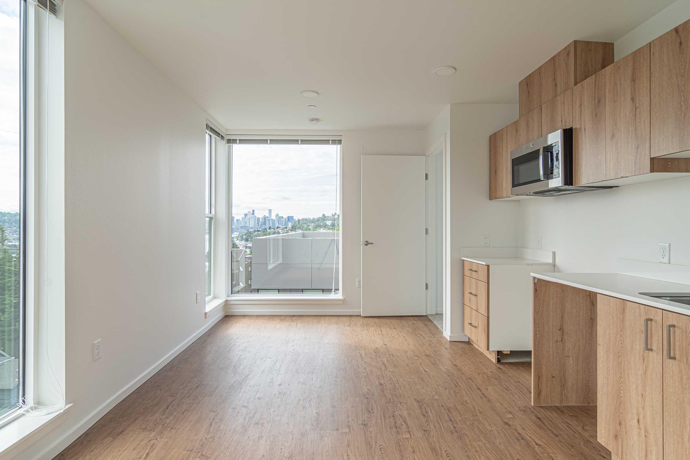 Modern, empty living space with large windows showcasing a city skyline. The room features light wooden floors, a small kitchenette with a microwave and cabinets, and a door leading to a balcony. Natural light fills the area, creating a bright and airy atmosphere.