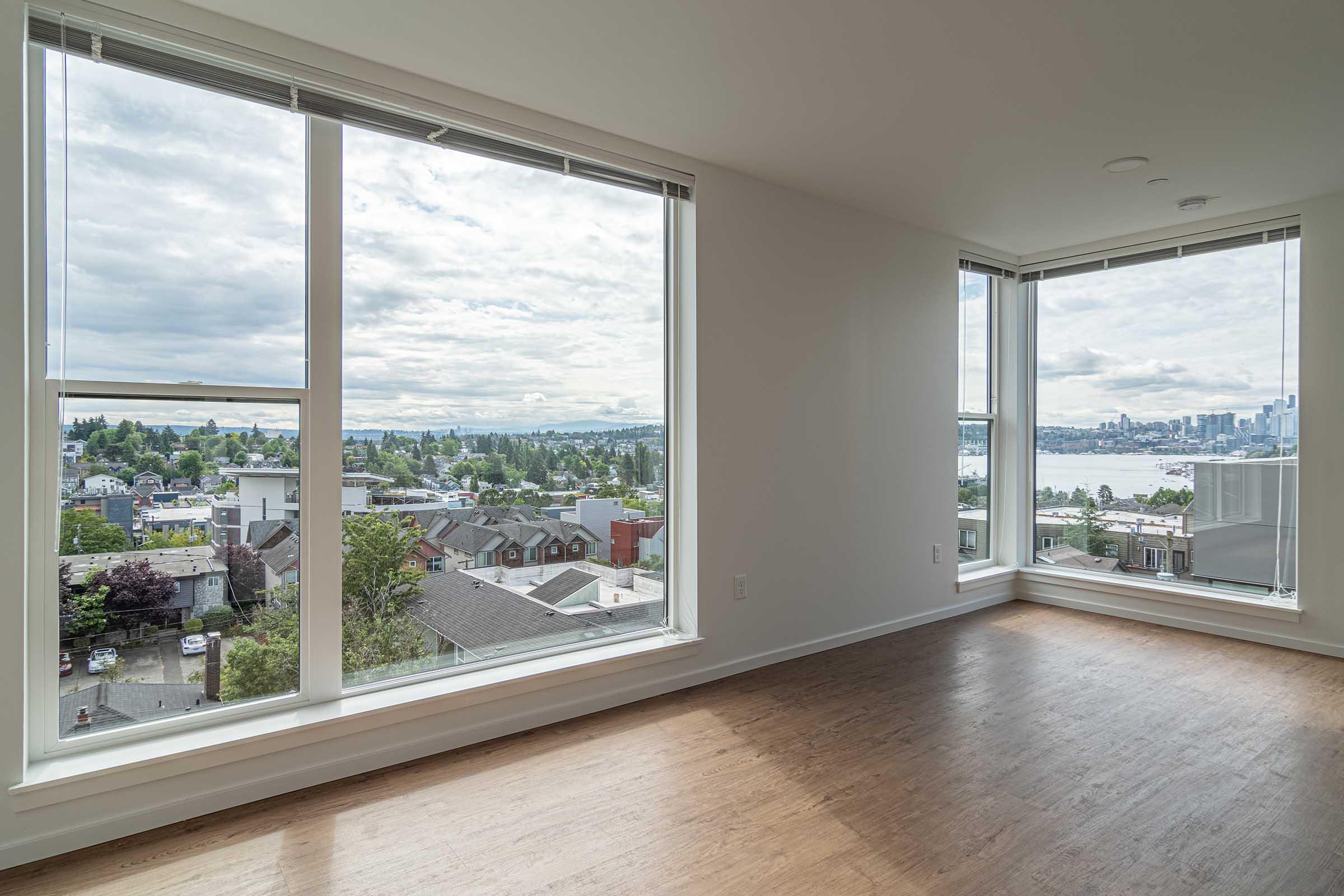 Bright and airy interior of a room with large windows providing a panoramic view of the city skyline and waterfront. Soft clouds fill the sky, while green trees and houses are visible in the foreground. The room features wooden floors and white walls, creating a modern and inviting atmosphere.