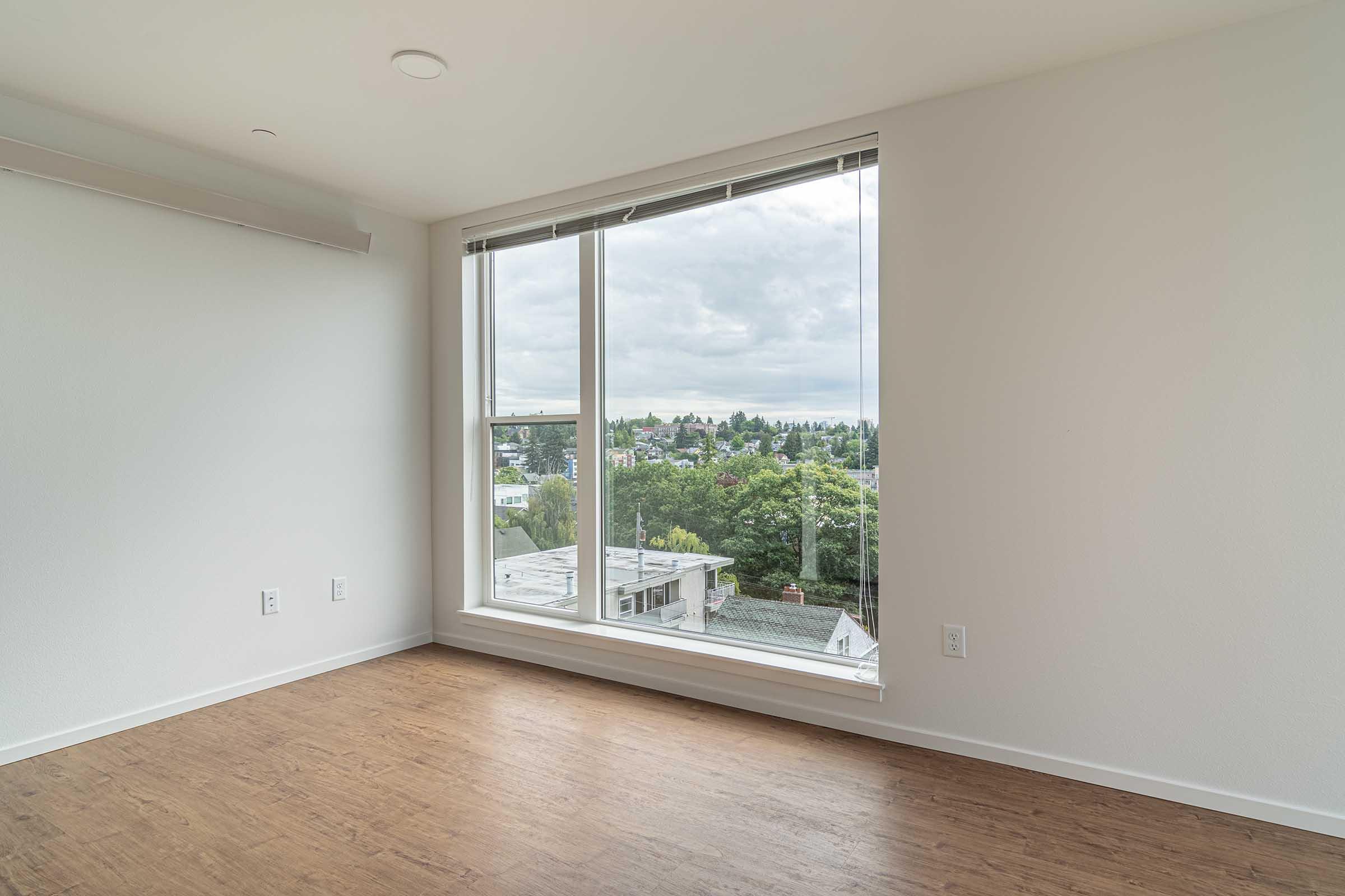 A bright, empty room with large windows offering a view of greenery and buildings outside. The walls are painted white, and the floor features wood-like laminate. Natural light illuminates the space, creating an inviting atmosphere.