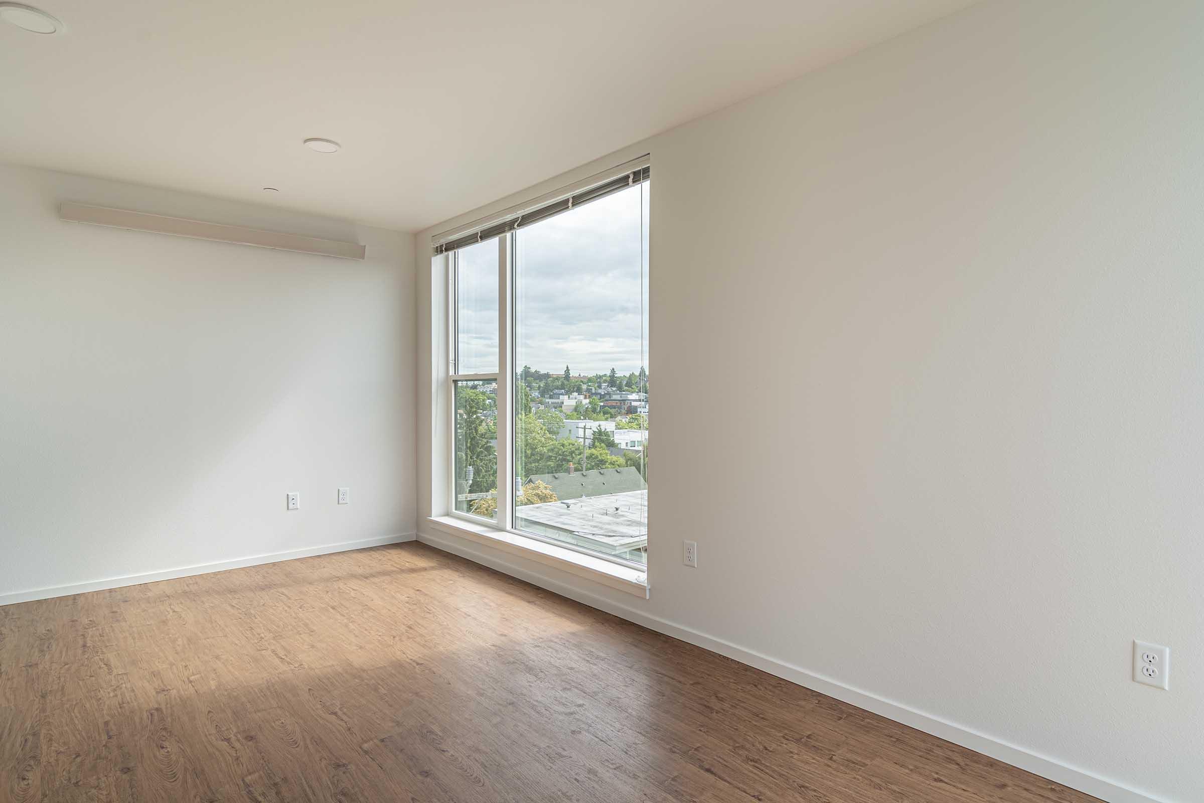 A bright and airy room featuring a large window with a view of the outdoors. The walls are painted in a light color, and the flooring is a warm wood tone. Natural light floods the space, creating an inviting atmosphere.