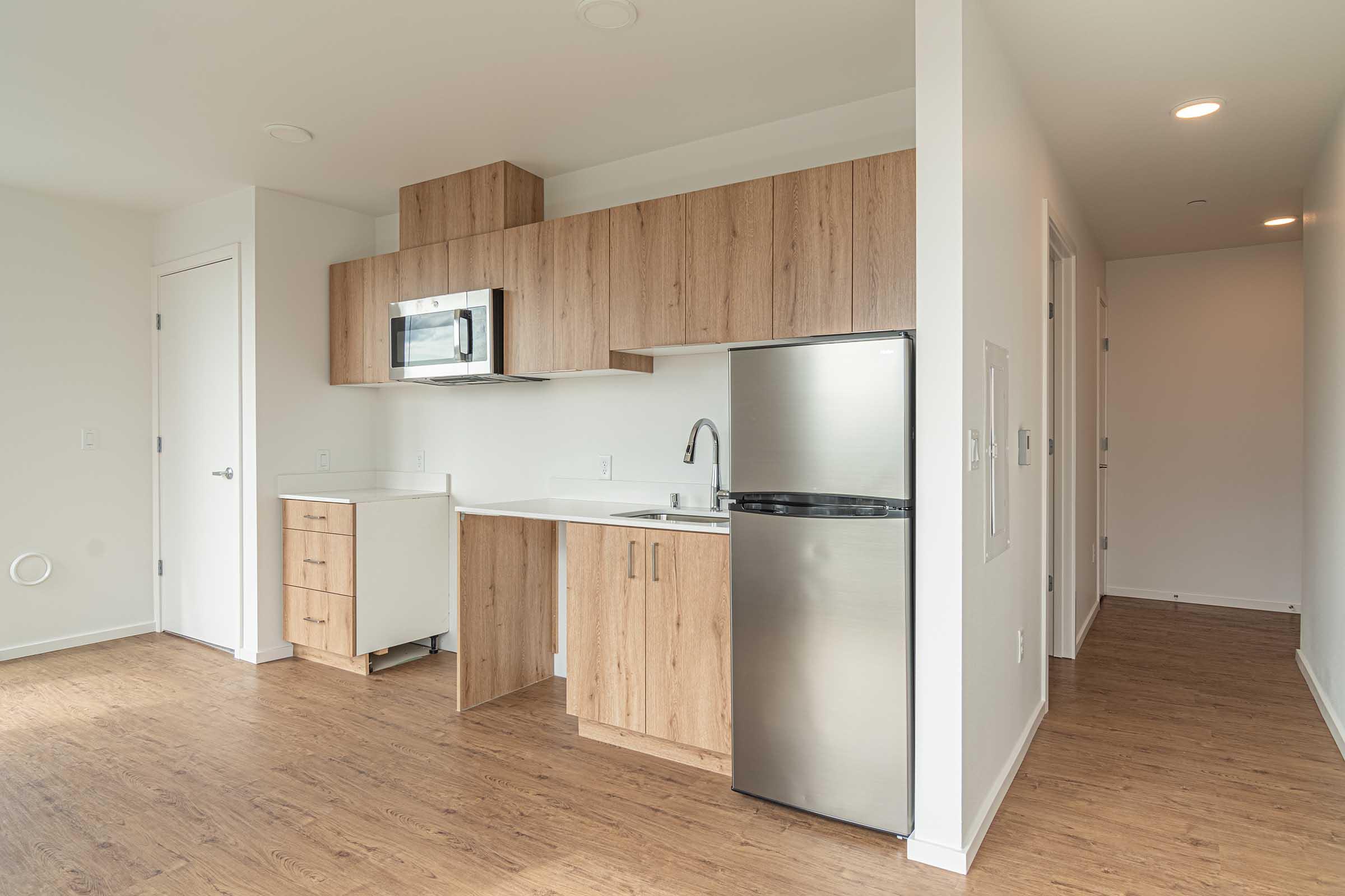 A modern kitchen featuring wooden cabinets, a stainless steel refrigerator, a microwave above the sink, and a minimalist design. The flooring is light wood, and the space has neutral-colored walls and simple fixtures, creating a clean and contemporary atmosphere. An open hallway is visible in the background.