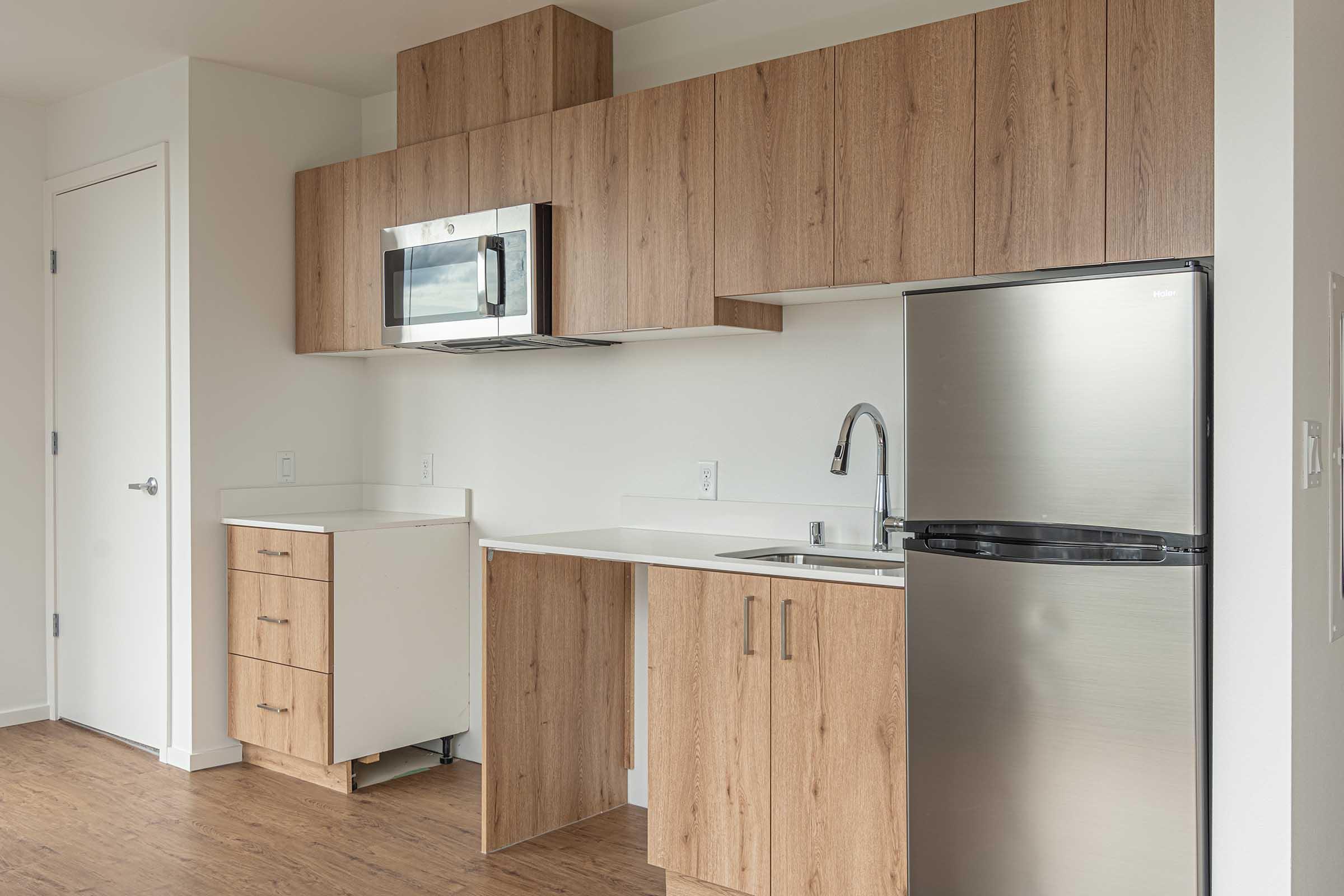 Modern kitchen in a light-colored space featuring wood cabinetry, a stainless steel refrigerator, a microwave mounted above the countertop, and a sink. The design is minimalist with a clean layout and neutral tones, providing an inviting atmosphere.