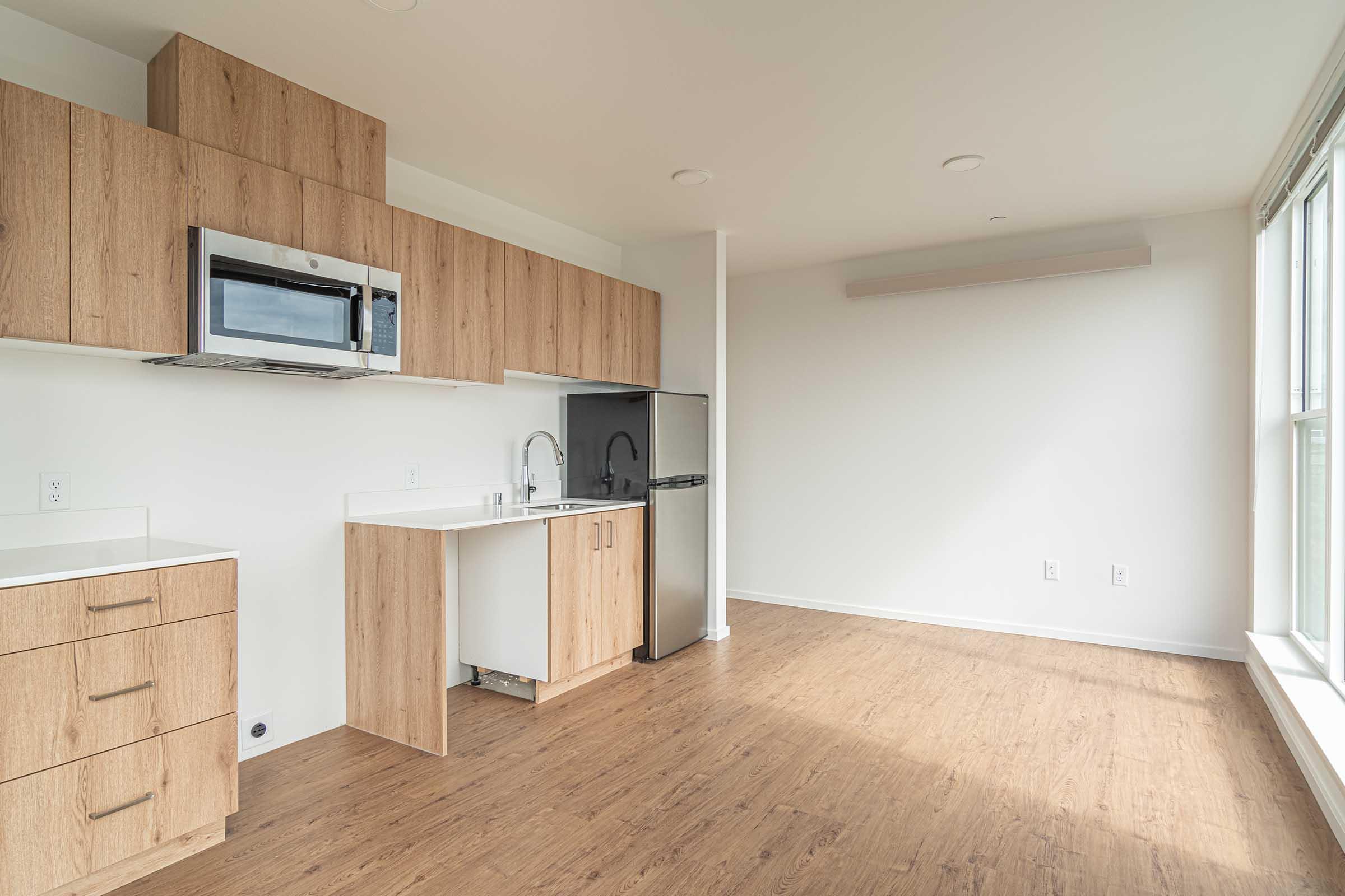 Modern kitchen area featuring wooden cabinets and drawers, a microwave above the countertop, a small sink with a faucet, and a black refrigerator. The space has light-colored walls and a large window allowing natural light. The flooring is a warm-toned laminate, creating a cozy atmosphere.