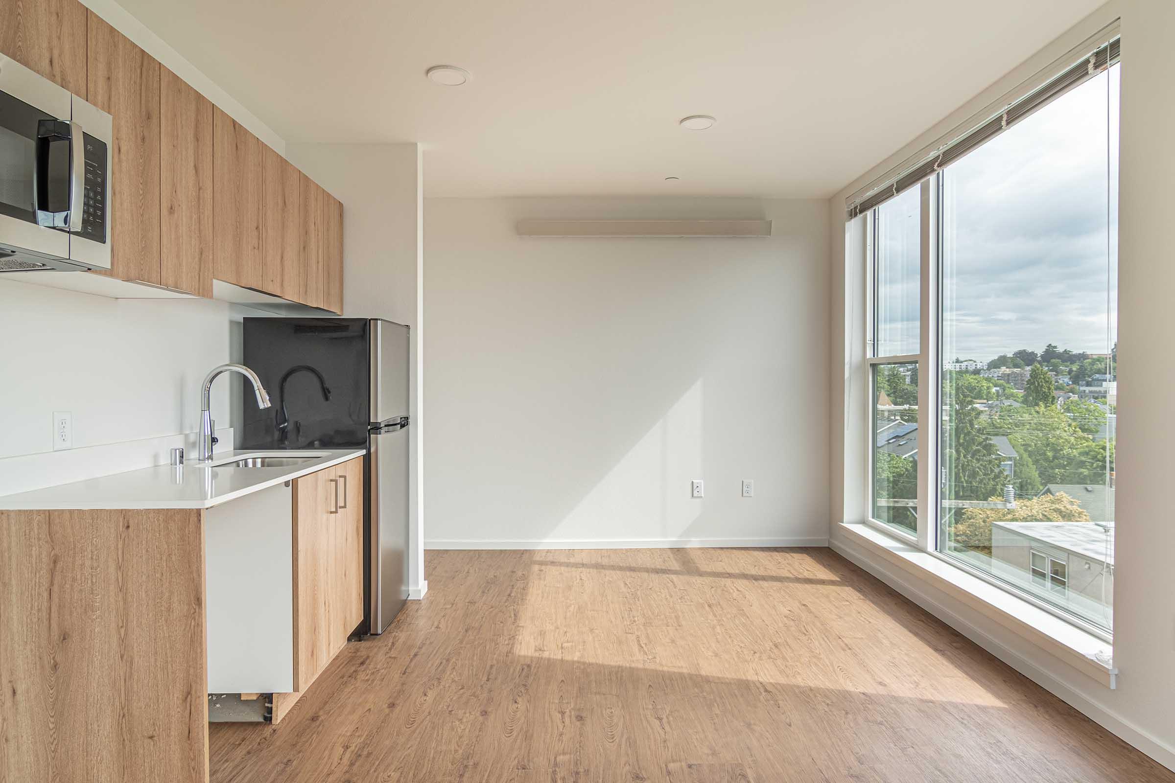 A modern, minimalist apartment interior featuring a kitchen area with wooden cabinets, a black refrigerator, and a stainless steel microwave. The room has large windows allowing natural light, a neutral-colored wall, and light wooden flooring, creating an airy and inviting space.