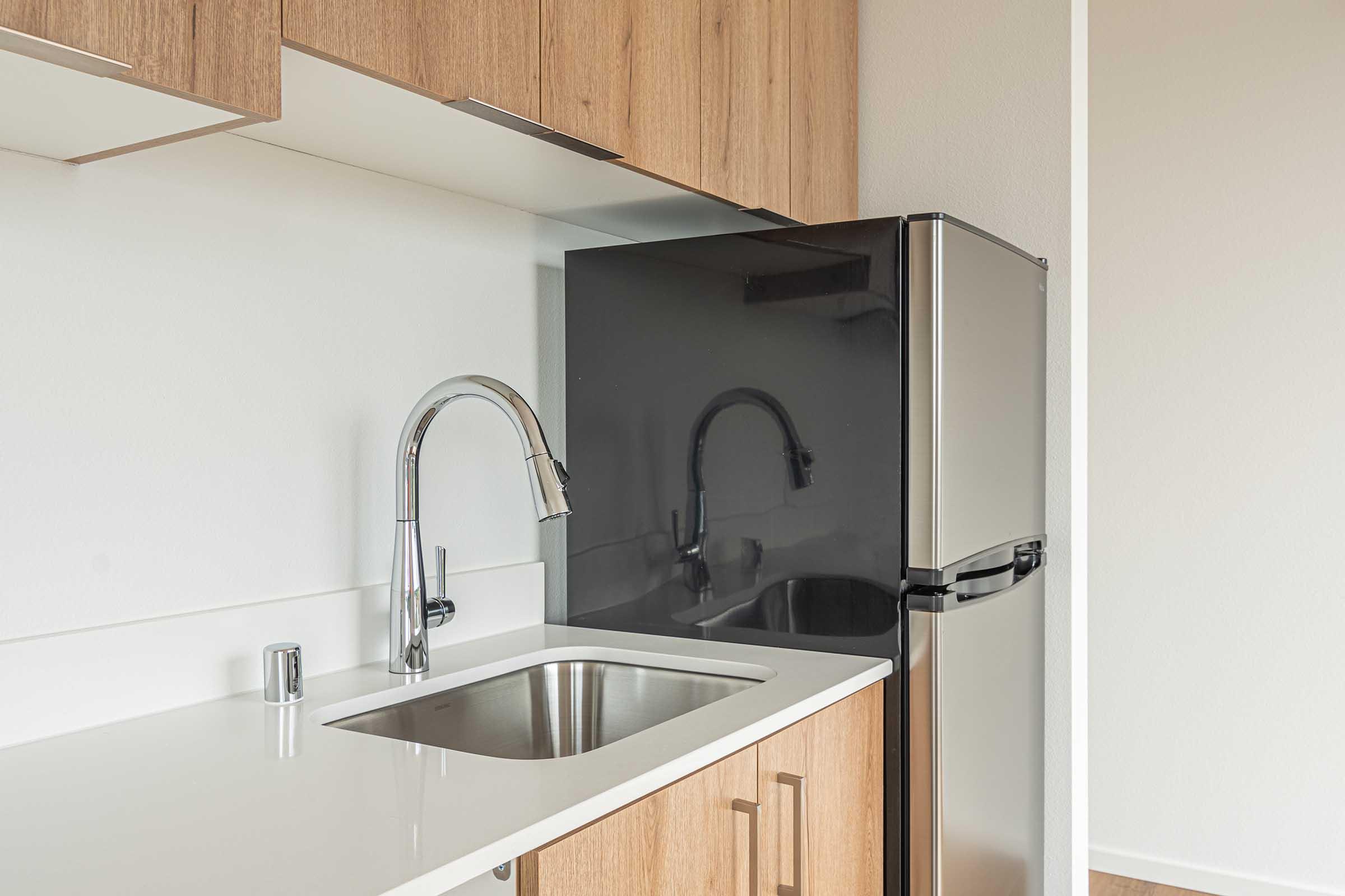 Modern kitchen interior featuring a sleek design with wooden cabinetry, a stainless steel sink with a stylish faucet, and a compact refrigerator. The countertop is smooth and white, complementing the cabinetry, while the walls are painted in a light shade, creating a bright and airy space.