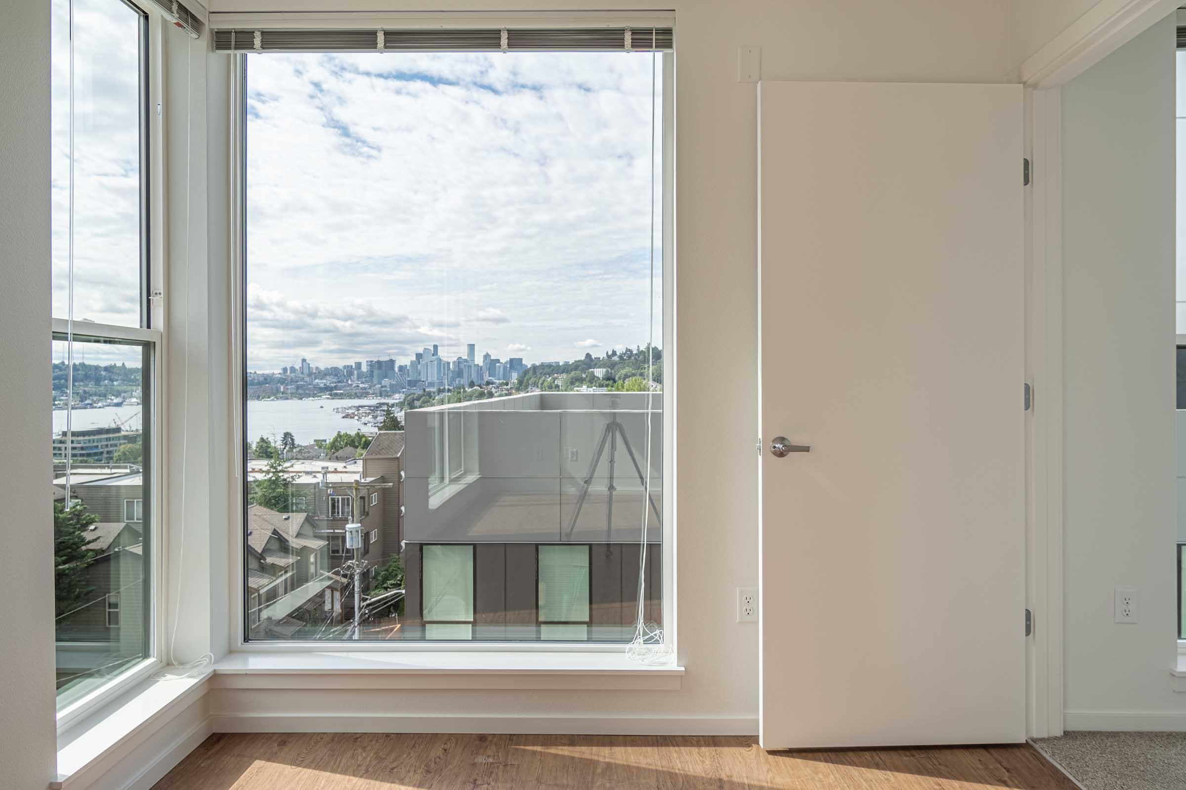 Interior view of a bright room showcasing a large window with a cityscape and water view outside. The window frames the Seattle skyline on the horizon, with partly cloudy skies above and a modern building in the foreground. A closed white door is visible on the right side of the image.