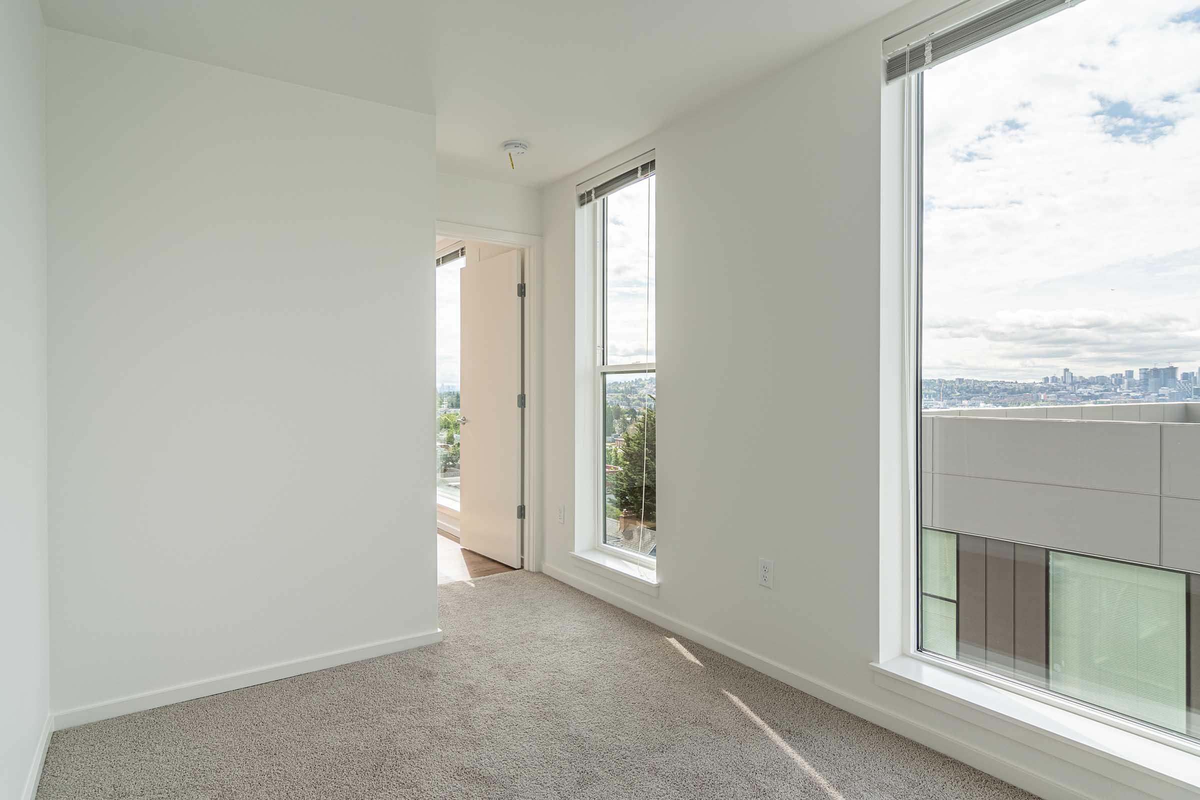 A bright, minimalist room with light-colored walls and large windows allowing natural light to enter. The floor is covered in soft carpet. A doorway leads to another area, and in the background, a city skyline can be seen through the windows. The overall look is modern and spacious.