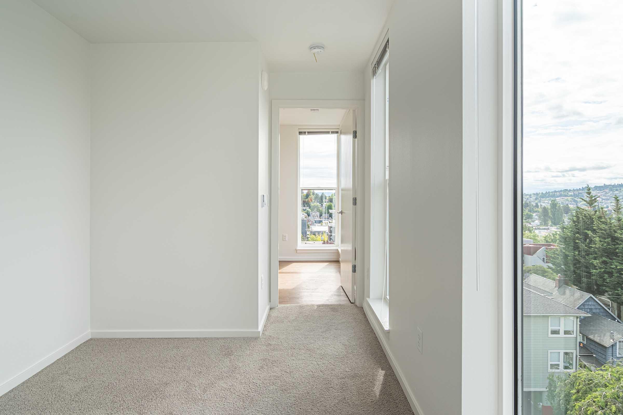 A well-lit, modern hallway leading to a window with a view. The walls are painted white, and the floor is carpeted in a light color. Sunlight enters from the end of the hallway, creating a welcoming atmosphere. The space appears clean and minimalistic, with no furniture present.