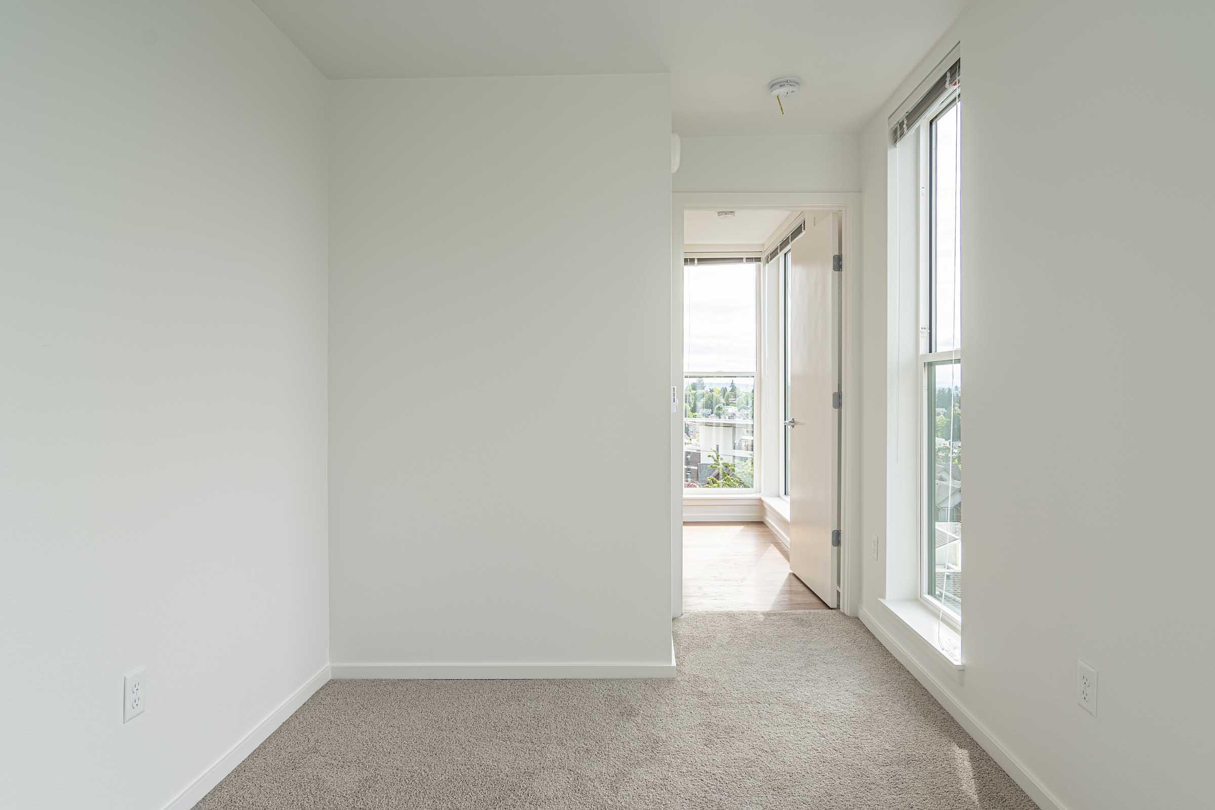 A bright, empty room with light gray walls and new carpet. Two large windows allow natural light to fill the space. A door leads to another area, visible in the background, with a view of the outdoors. The atmosphere is clean and minimalist, ideal for various uses such as a bedroom or office.