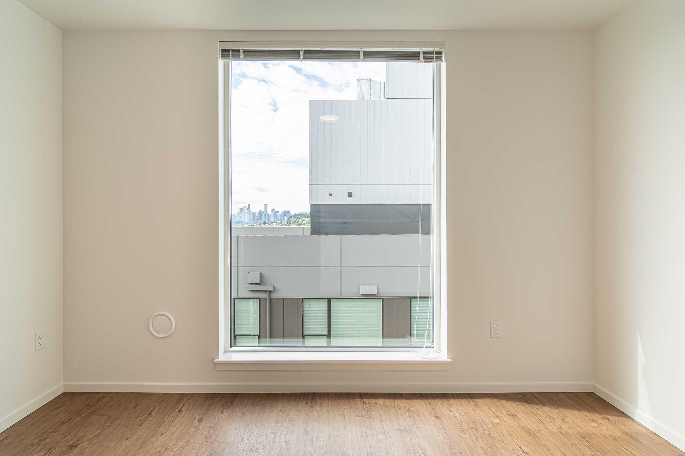 A bright and empty room with light-colored walls and a large rectangular window. The window showcases a view of a city skyline in the background, framed by modern building exteriors. The floor is wood-like, adding warmth to the minimalist space.